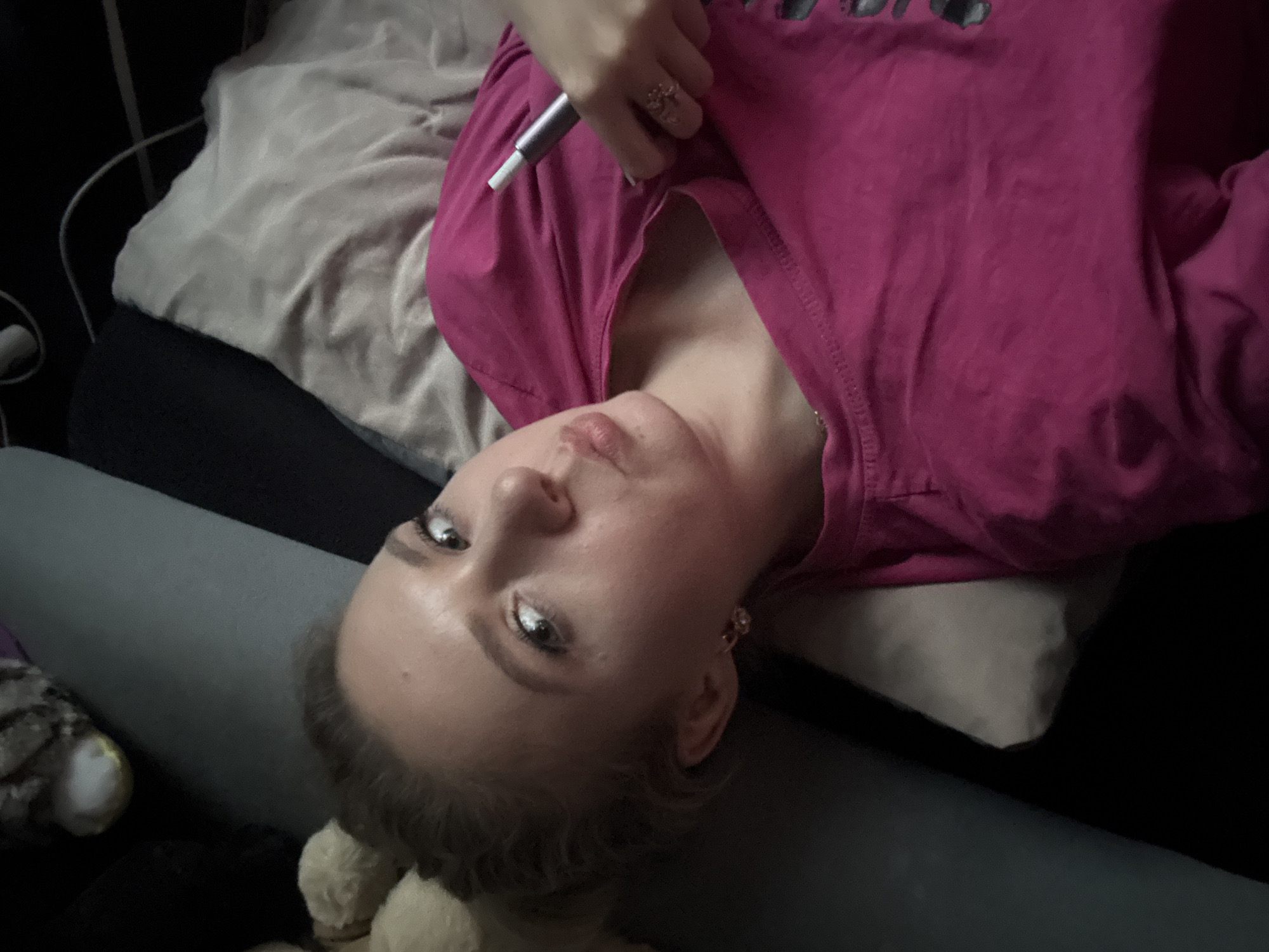 In an indoor setting, a Caucasian female with blonde hair is lying down, holding a gray electronic cigarette device in her right hand. She is wearing a pink t-shirt. Her head is tilted and she is looking towards the camera with her lips slightly pursed. The background consists of pillows and what appears to be a dark couch. There are no discernible elements to indicate a specific location within a city.