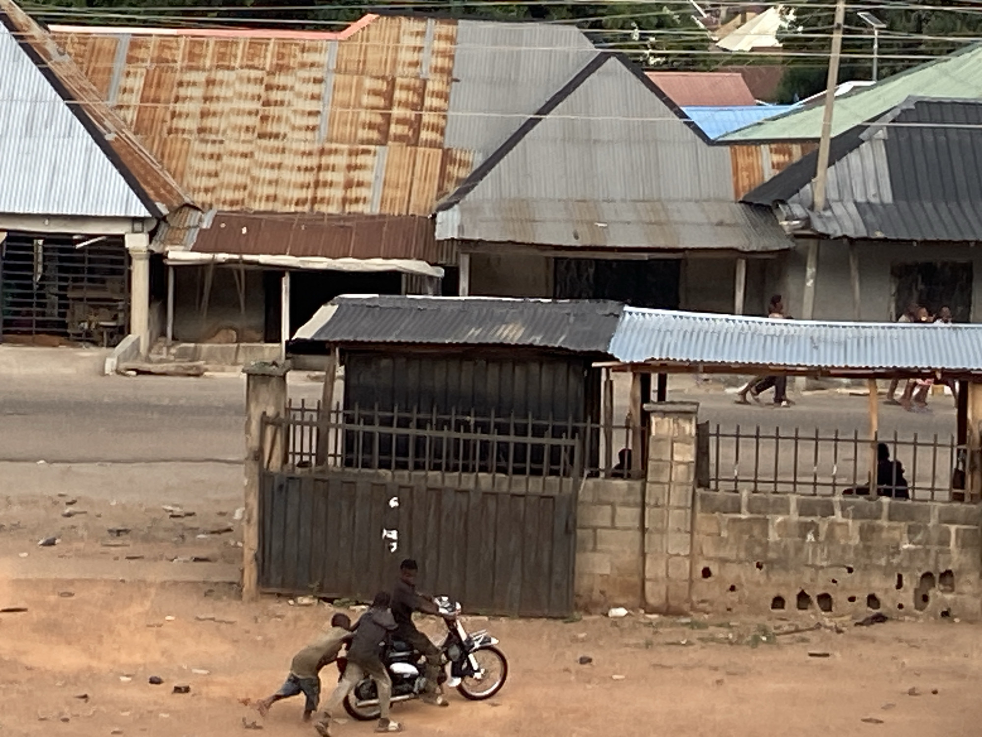 The image depicts a street scene in Tudun Wada Bibinu, Nigeria. In the foreground, three young boys are interacting with a motorcycle. One boy is seated on the motorcycle, while the other two are on either side, seemingly pushing or helping to maneuver the vehicle. The background shows several single-story buildings with corrugated metal roofs of varying colors and conditions, some appearing rusted. Pedestrians are visible walking on the street in the middle ground. A fence constructed of stone blocks and metal bars is situated between the children and the street. The area appears to be a residential neighborhood.