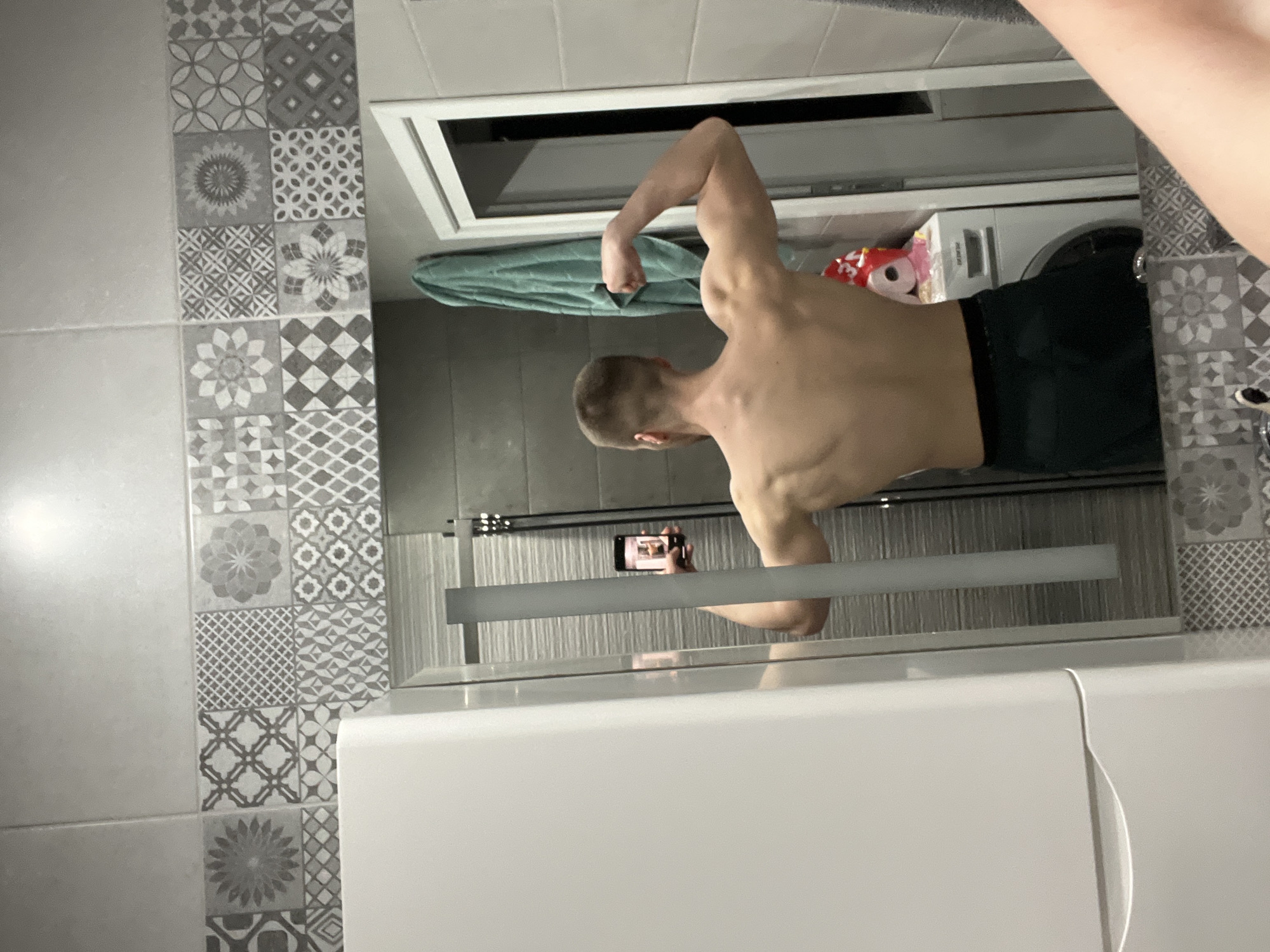 The image depicts a person taking a mirror selfie in a bathroom. The subject is a shirtless man, seen from the back in the mirror's reflection, flexing his left bicep. He holds a black smartphone horizontally to capture the image.
The bathroom features a wall adorned with decorative grey and white patterned tiles, arranged in a grid of various geometric and floral designs. A plain light grey tiled wall is visible to the left. A clear glass shower partition, with a frosted horizontal bar across its middle, separates the shower area from the rest of the bathroom, and the man's reflection is seen through this partition.
In the background reflection, behind the man, a white washing machine is visible, with the brand name "CANDY" faintly discernible on its front panel. A folded green towel rests on top of the washing machine, and other items, including what appears to be a red-packaged product, are on a shelf above it. The white surface of a bathtub or vanity unit occupies the foreground. The image is brightly lit, suggesting indoor lighting, and the time of day is not discernible. The location context provided for this image is Burgas, Bulgaria.
