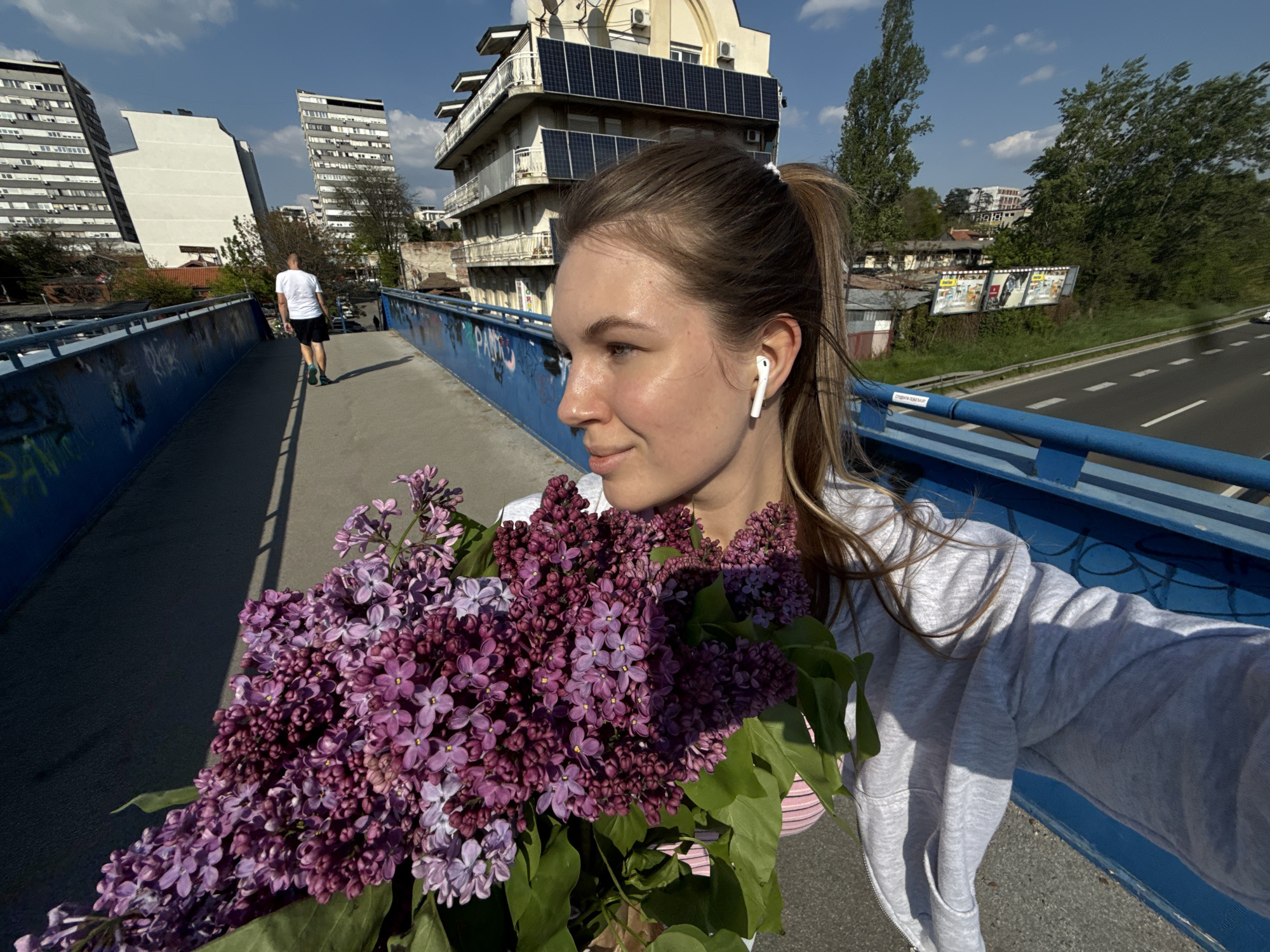 A young woman is captured in a selfie on an elevated pedestrian bridge in Vozdovac Urban Municipality, Serbia, holding a substantial bouquet of purple lilacs. She is seen in profile, looking slightly to her left with a serene expression, her light brown hair tied in a ponytail, and a white earbud in her right ear. She wears a light grey zip-up hoodie.
The large bouquet of vibrant dark and light purple lilacs, interspersed with green leaves, occupies the left foreground, basked in strong sunlight that casts clear shadows.
In the mid-ground, a man in a white t-shirt and dark shorts walks away from the camera along the bridge. The bridge features a prominent blue railing with some graffiti.
The background depicts a sunny urban landscape. To the left, tall, grey residential buildings stand against a clear blue sky with sparse clouds. To the right, another multi-story building is visible, notably featuring numerous solar panels on its facade and roof. Beyond the bridge's railing, a multi-lane road with a visible white dashed line runs parallel, flanked by trees and billboards. The scene suggests a bright, pleasant day in an urban setting.
Visible text includes illegible graffiti on the blue bridge railing and indistinct text on distant billboards.