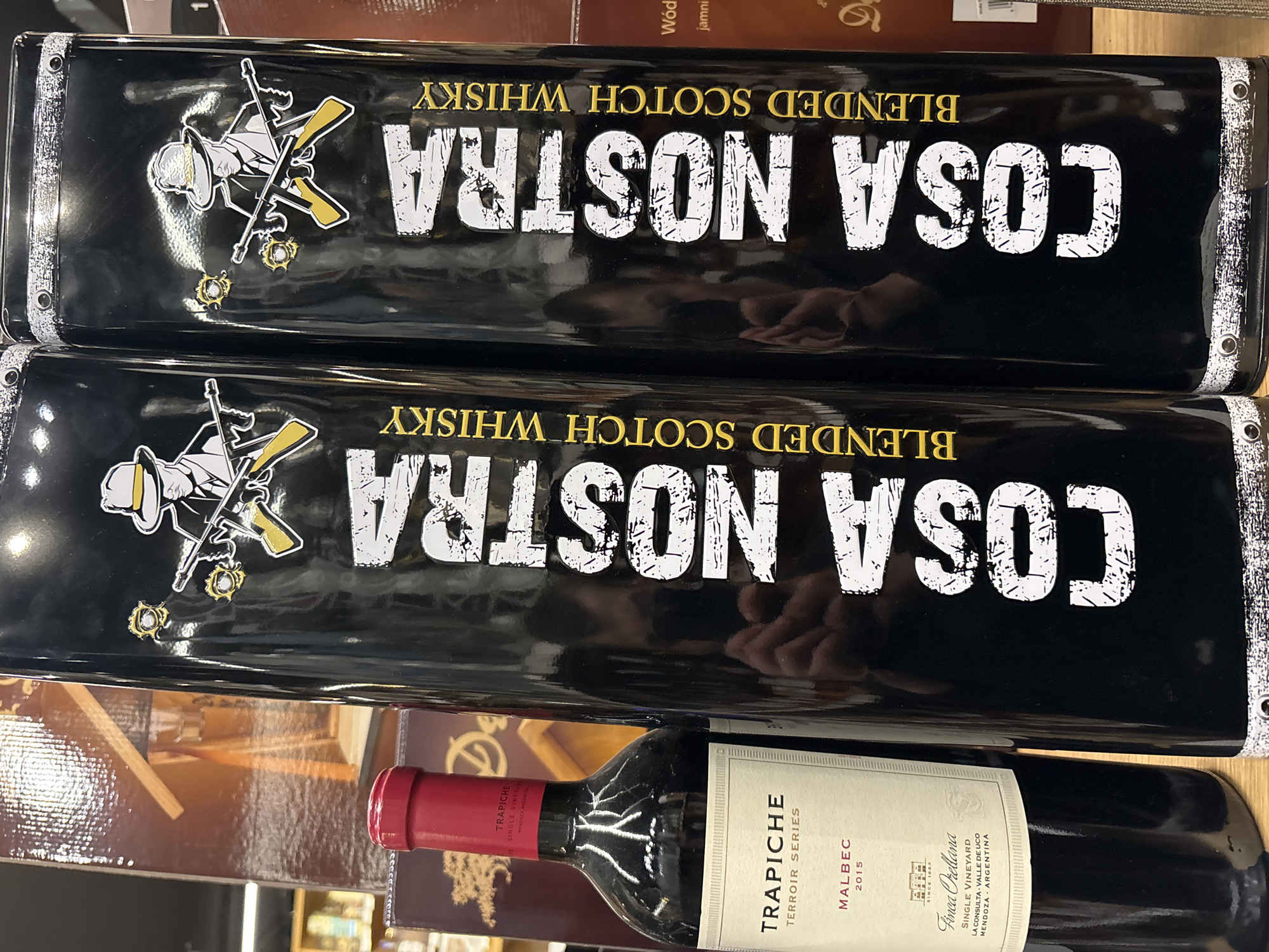 Here is a detailed description of the image:
The image shows two black containers stacked on top of each other, with the words "COSA NOSTRA" printed in large white letters and "BLENDED SCOTCH WHISKY" in smaller golden letters. There is also a logo of a gangster figure with guns and bullet holes. Underneath the containers is a wine bottle labeled "TRAPICHE TERROIR SERIES MALBEC 2015," including details about the vineyard and its location in Mendoza, Argentina. The setting appears to be indoors, possibly a store or shelf. There is no indication of the city of Plovdiv, Bulgaria, in the image.