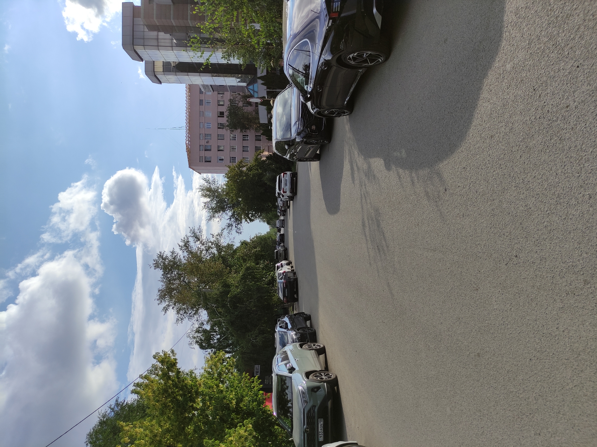 The image shows a street scene in the city of Semey, Kazakhstan. A line of cars is parked along the right side of the asphalt road. In the foreground, a dark-colored sedan is parked, and to its left, a black SUV is visible. Further down the road, more cars are parked, including white SUVs and sedans. On the left side of the road, trees line the sidewalk, and a light-colored SUV is parked. In the background, modern buildings, including a multi-story building with glass windows, are visible. The sky is blue with scattered white clouds, and the sun is casting shadows on the road, indicating it is likely late afternoon. The overall impression is of a typical urban street with parked vehicles.