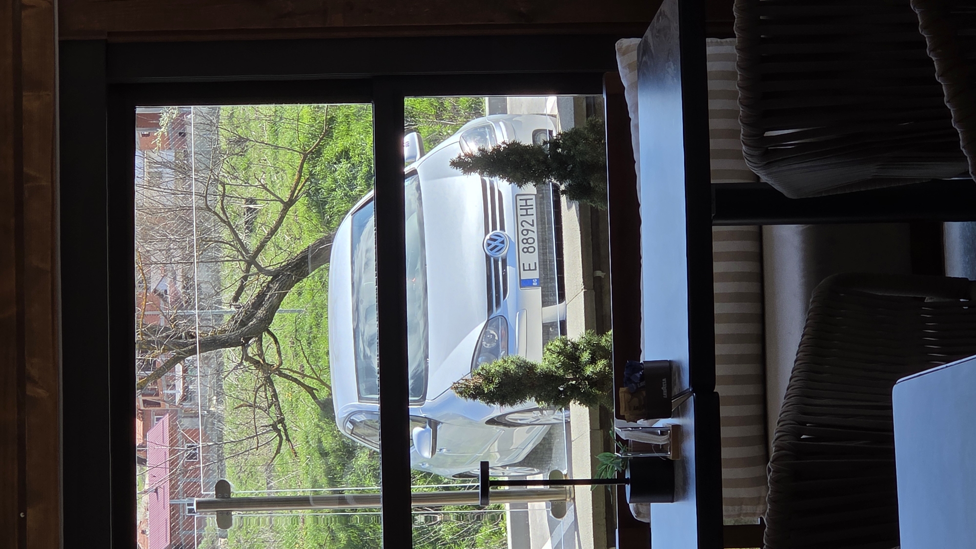 The image captures a view through a window, offering a glimpse of the outside. A white Volkswagen car with the license plate "E 8892 HH" is parked outside. The surrounding environment includes greenery and a tree. In the foreground, inside the room, parts of chairs and a table are visible. There are some items on the table near the window. Based on the license plate, the location is in Bulgaria, but further specifics about the city or region are not discernible from the image.