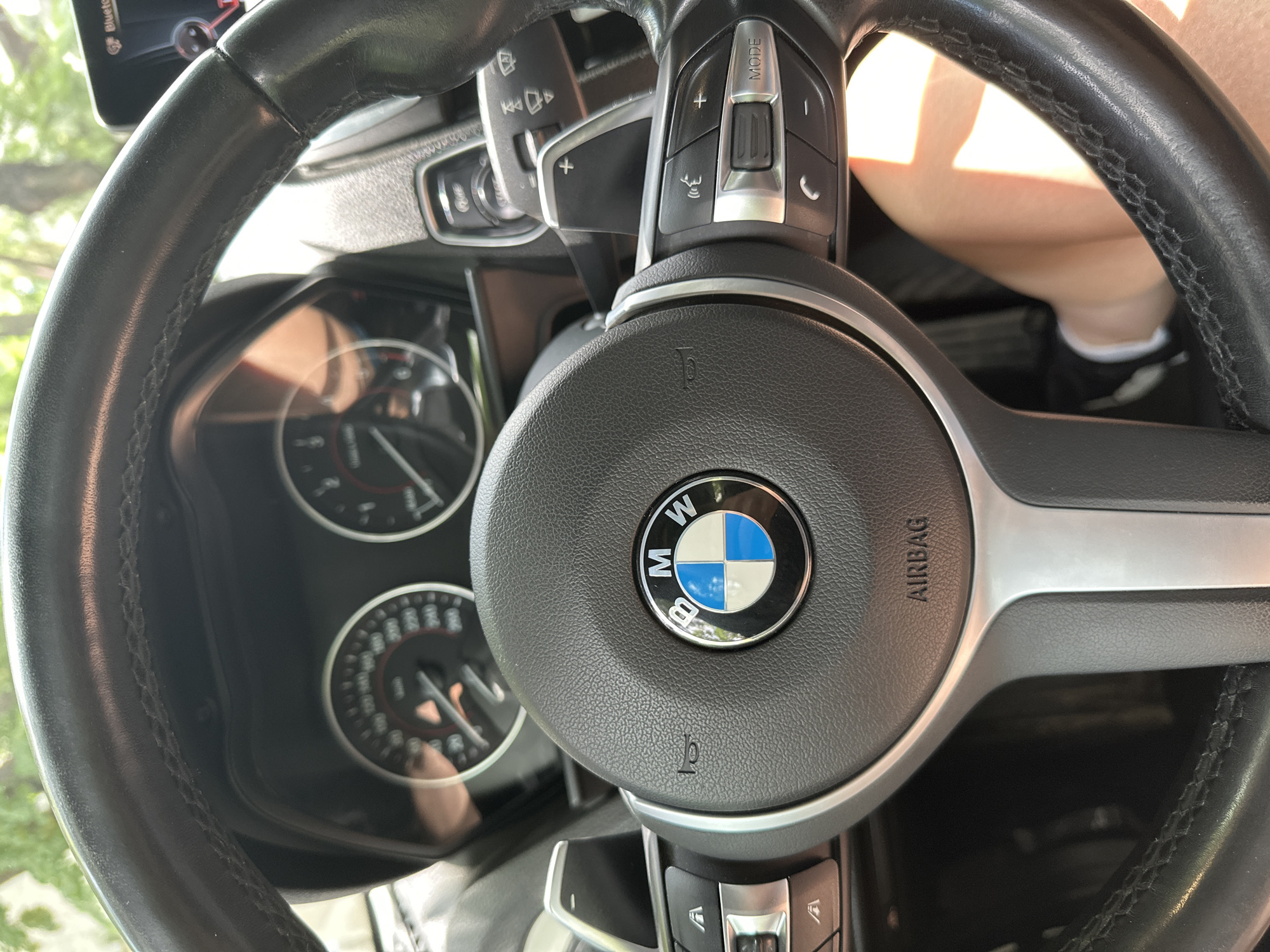 The image shows a close-up view of the steering wheel and dashboard of a BMW car, as seen from the driver's seat. The BMW logo is prominently displayed in the center of the steering wheel. The dashboard features two analog gauges, likely for speed and RPM, with a digital display between them. There are also various buttons and controls on the steering wheel, including paddle shifters and buttons for audio and phone functions. A person's leg and foot are partially visible in the lower right portion of the image, indicating the driver is seated inside the car. The background is blurred, but appears to be outdoors with trees visible, suggesting a sunny day. There is no indication of the specific location within the city of Sofia, Bulgaria.