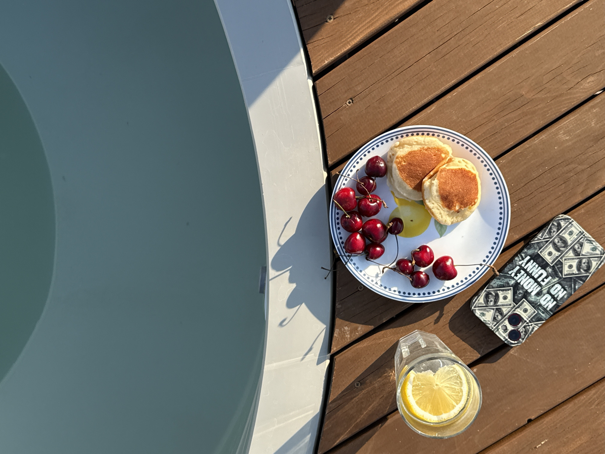 A plate of pancakes and cherries sits on a wooden deck next to a glass of water with a lemon slice. A phone case with a money pattern is also on the deck. In the background, the edge of a body of water is visible. The scene is outdoors, likely in a residential area. No people are visible. The location is Starokorsunskaya, Russia.