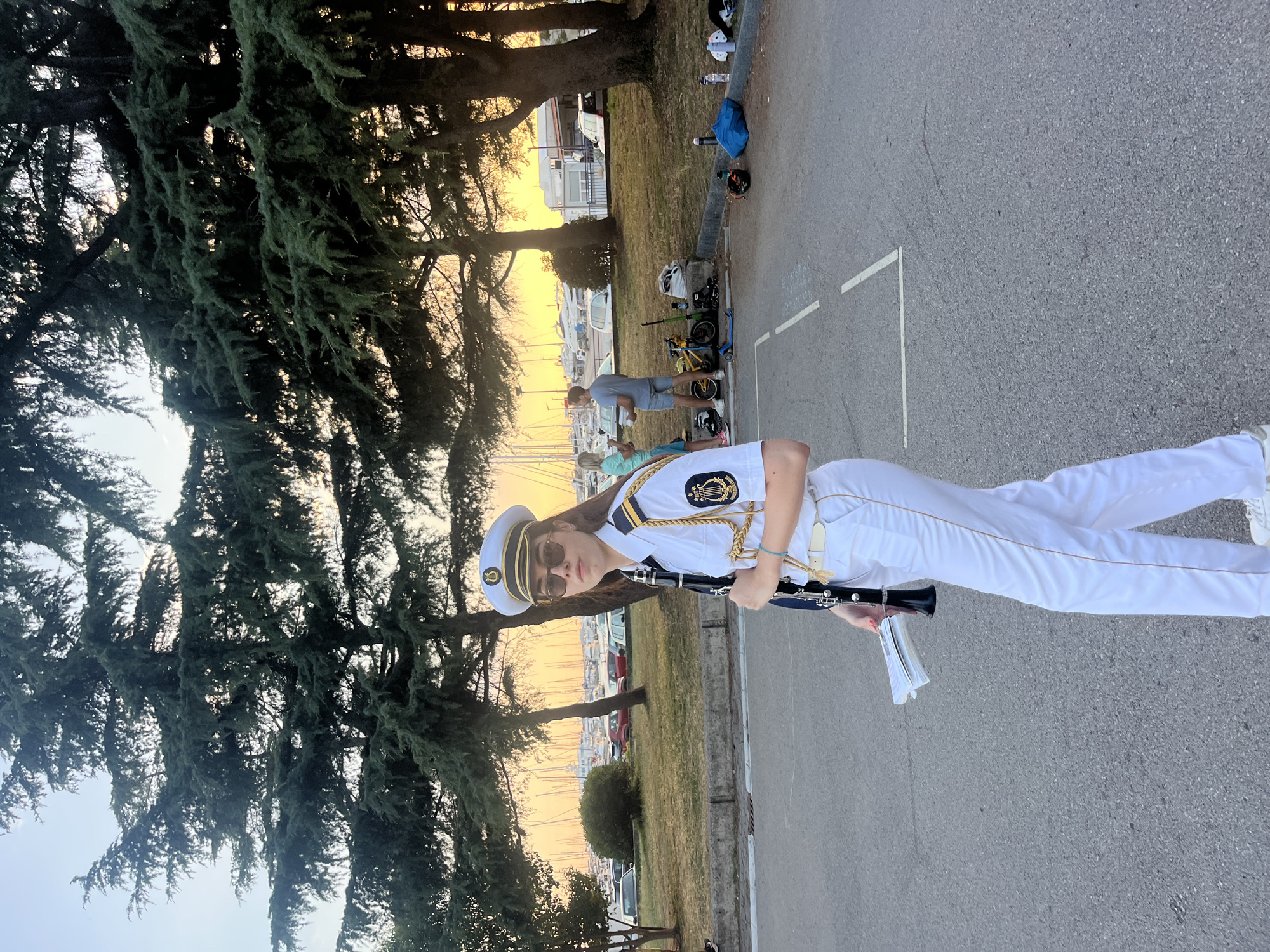 A young woman, dressed in a white naval-style uniform with gold trim and a white cap, stands in a paved area with a clarinet and music sheets. She is wearing sunglasses and her gaze is directed towards the camera. In the background, a marina with numerous boats is visible, along with trees and a few other people in casual attire. The scene appears to be set during sunset, with the warm glow of the setting sun filtering through the trees. The location is identified as Šušanj, Montenegro.