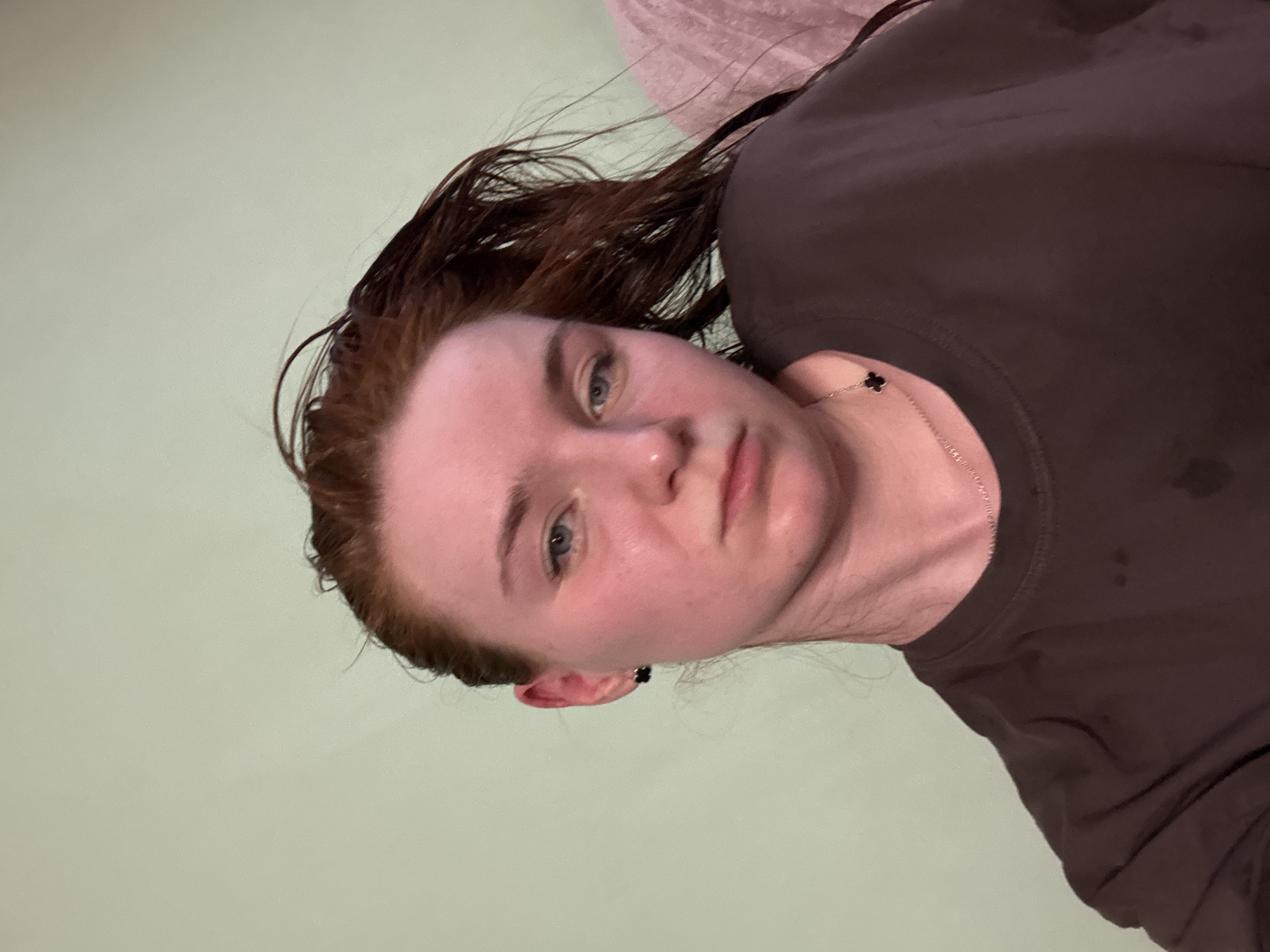 A close-up, slightly rotated, selfie of a person with wet, reddish-brown hair. The person has fair skin and blue eyes. They are wearing a dark brown shirt and a delicate silver necklace with a black clover pendant. The background is a light green wall, and part of a pink object is visible in the top-right corner. The scene appears to be indoors. The location is specified as Unknown, Russia, but there is no discernible evidence in the image to confirm this or any specific location.