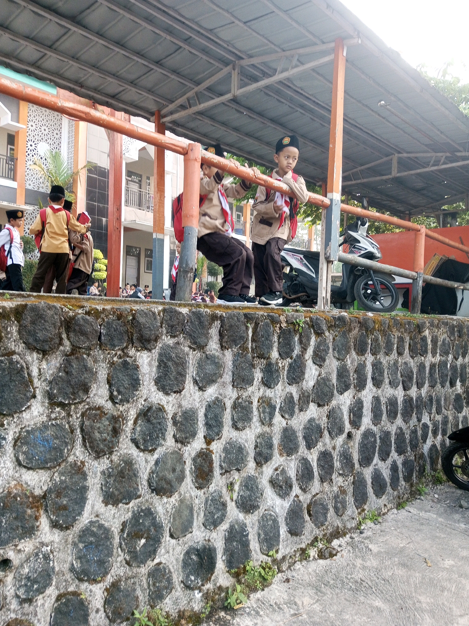 In Krasak, Indonesia, several young students in scout uniforms are gathered on an elevated platform with a covered roof. Two boys, wearing tan uniforms with red scarves and backpacks, are the most prominent figures. One is perched on a railing, looking down, while the other stands next to him. In the background, more students are visible, some seated and others standing. A motorcycle is parked to the right, and the scene is bordered by a stone wall in the foreground. The architectural style of the building behind the students suggests a school or community center. The lighting indicates it is daytime, likely with sunlight filtering through the overcast sky.