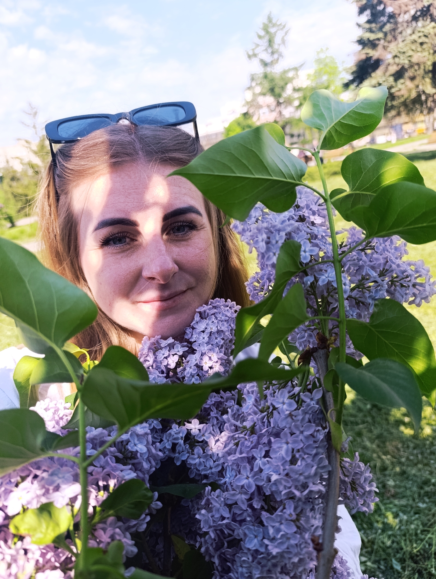 A woman with light brown hair pulled back with sunglasses on her head holds a branch of purple lilac flowers. The woman's face is partially shadowed by the flowers and leaves. The background appears to be a park with trees and grass, suggesting an outdoor setting. The location is in a city, but the specific city is not known.