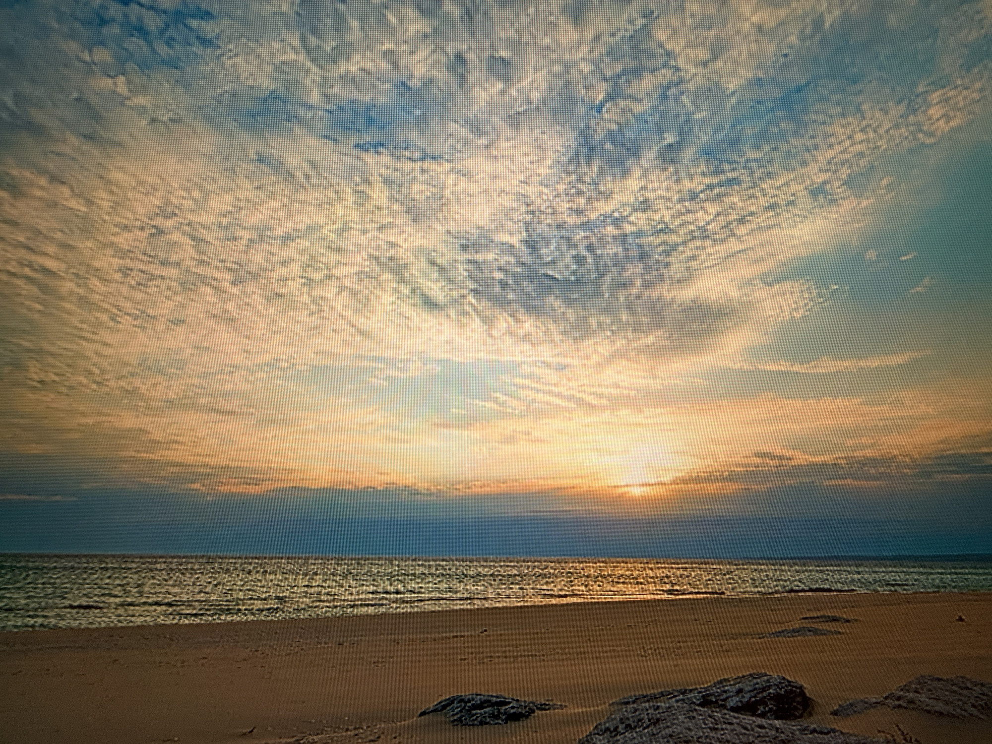 This image depicts a serene beach scene at sunset. The sky is filled with dramatic, wispy clouds illuminated by the warm hues of the setting sun, ranging from pale yellow to soft orange and pink. The sun itself is partially obscured by a band of darker clouds near the horizon, creating a sense of diffused light. Below the sky, a calm sea stretches out, its surface rippling and reflecting the golden light of the sunset. The foreground is a sandy beach, with a few scattered rocks and pebbles. The overall mood is peaceful and tranquil, evoking the feeling of a quiet evening by the sea. There is no visible text in the image. The location context of Szczecin, Poland, suggests this could be a coastal area near the Baltic Sea.
