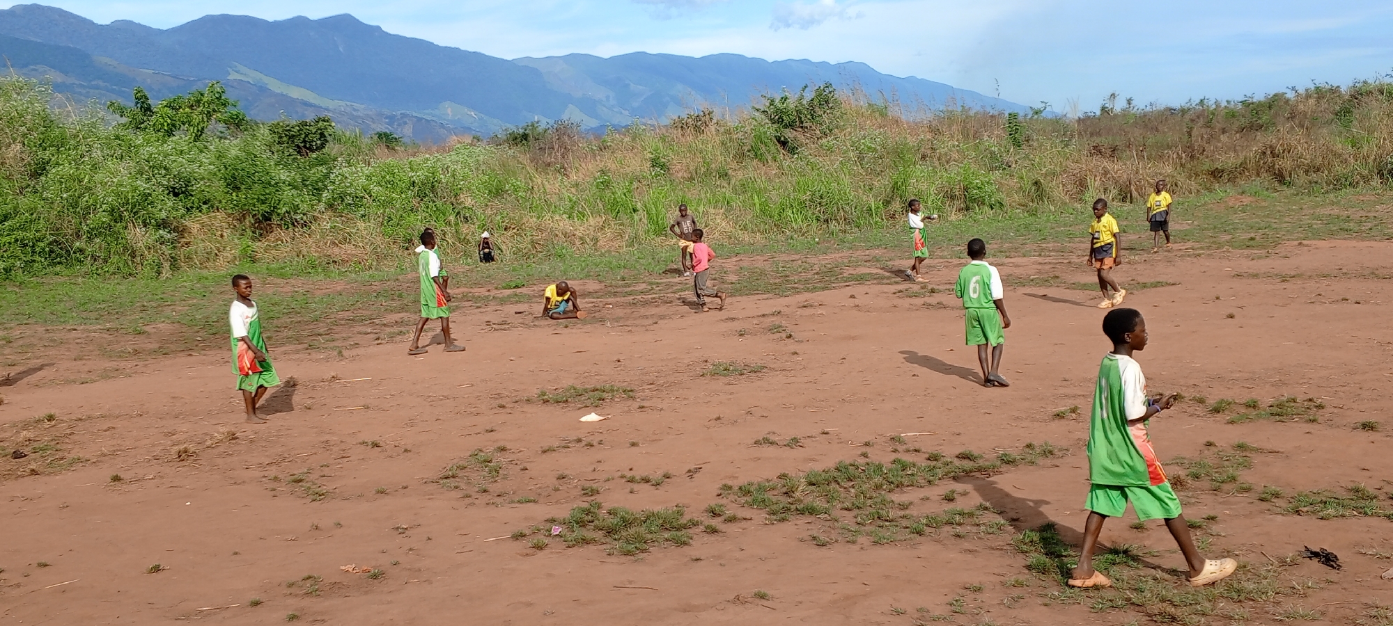 The image depicts a group of children playing on a dirt field in Ngalula, Democratic Republic of the Congo. In the background, there are visible mountains. Ten children are seen in the photo. Some are wearing green and white soccer uniforms, while others are wearing yellow and blue shirts. One child is wearing a pink shirt. One child is on the ground. The field is mostly dirt, with some scattered patches of grass. Trees and vegetation are visible in the background, behind the field. The sky is mostly clear with some clouds.