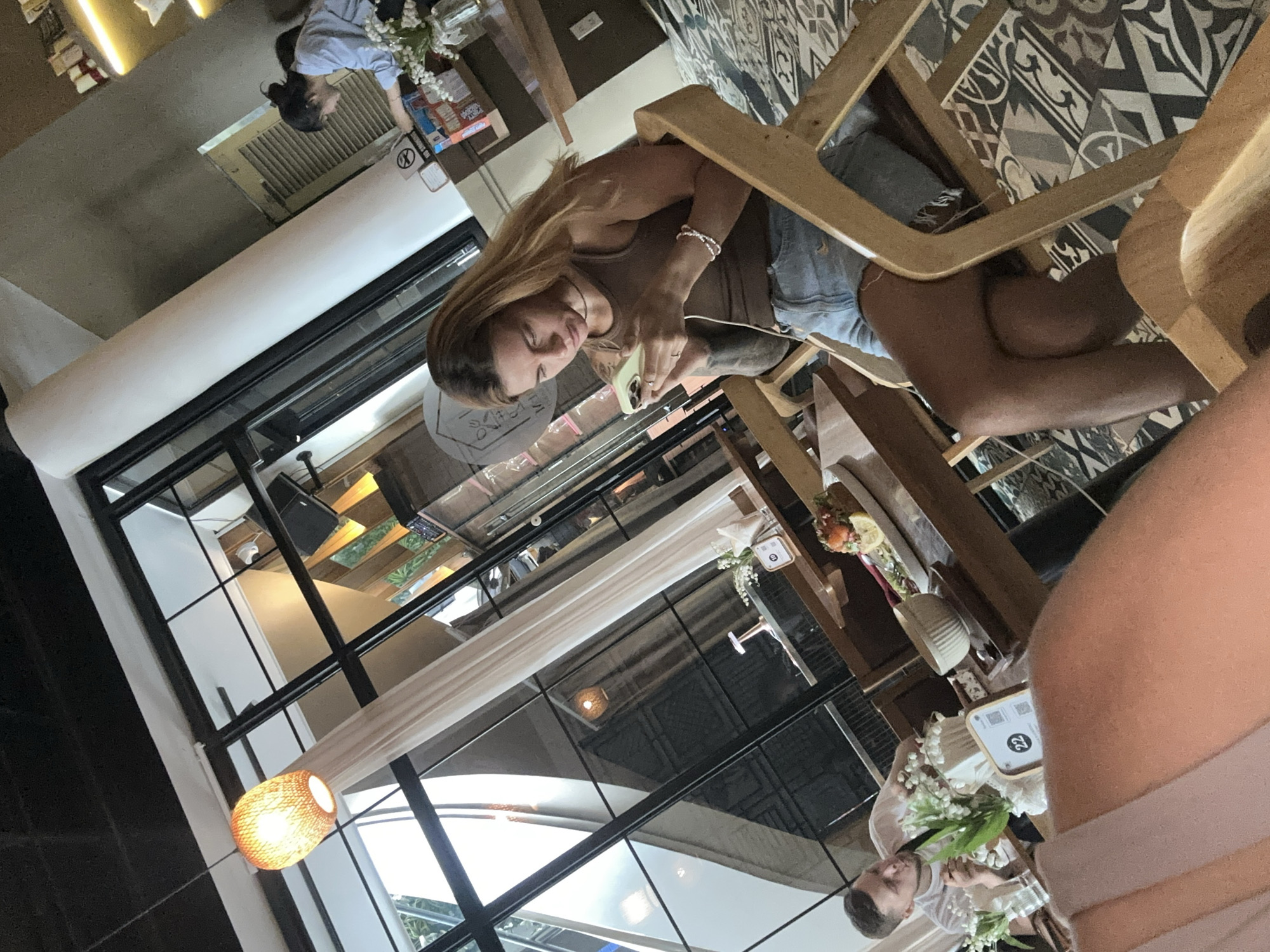 The image depicts the interior of a building in Nha Trang Ward, Vietnam. A woman with long brown hair sits at a wooden table, using a smartphone. She wears a tank top and denim shorts. A second person is partially visible on the right side, wearing a light-colored top. The table is decorated with flowers and a table number card. The building features large windows and a glass roof, through which light enters. A decorative wicker lamp hangs near the window. The building's interior design incorporates geometric patterned tiles on the floor.