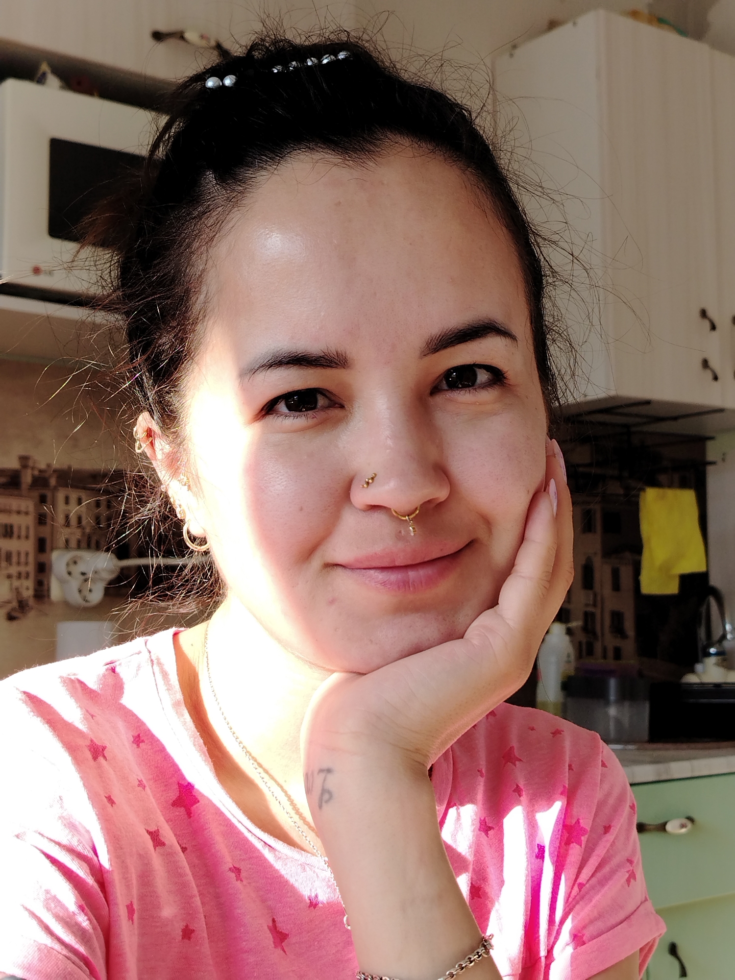A fair-skinned woman with dark hair pulled back in a bun is posing indoors. Her right hand is propping up her chin. She is wearing a pink t-shirt with star patterns, a gold nose ring, gold earrings, a gold necklace, and a gold bracelet on her left wrist. A faint tattoo is visible on her left forearm. The background shows a kitchen with white cabinets, a microwave, and other kitchen objects. The presence of buildings on the backsplash suggests Noviy, Russia, as this city is known for architectural themes incorporated in home interiors.