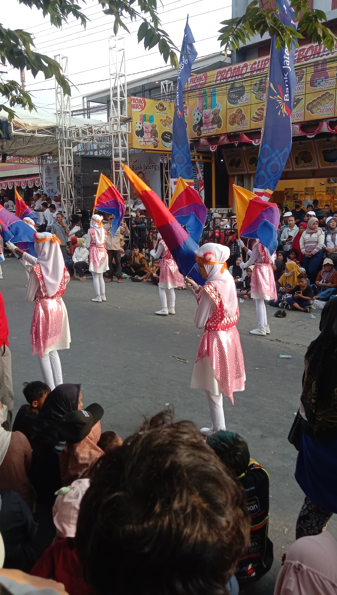 The image captures a daytime outdoor scene, likely in Jatisawit, Indonesia, featuring a parade or performance. Several young people, predominantly female and dressed in white outfits with pink, sparkling accents and head coverings, are holding large, colorful flags with purple, red, yellow, and blue sections. They appear to be performing a synchronized routine. In the background, a crowd of spectators is seated and standing, watching the event. Booths with promotional banners, including one advertising "PROMO HEBOH" and "Bank Jateng," are visible behind the performers. The environment includes a paved ground, temporary stage structures, and overhead tree branches. The overall atmosphere suggests a community event or festival.