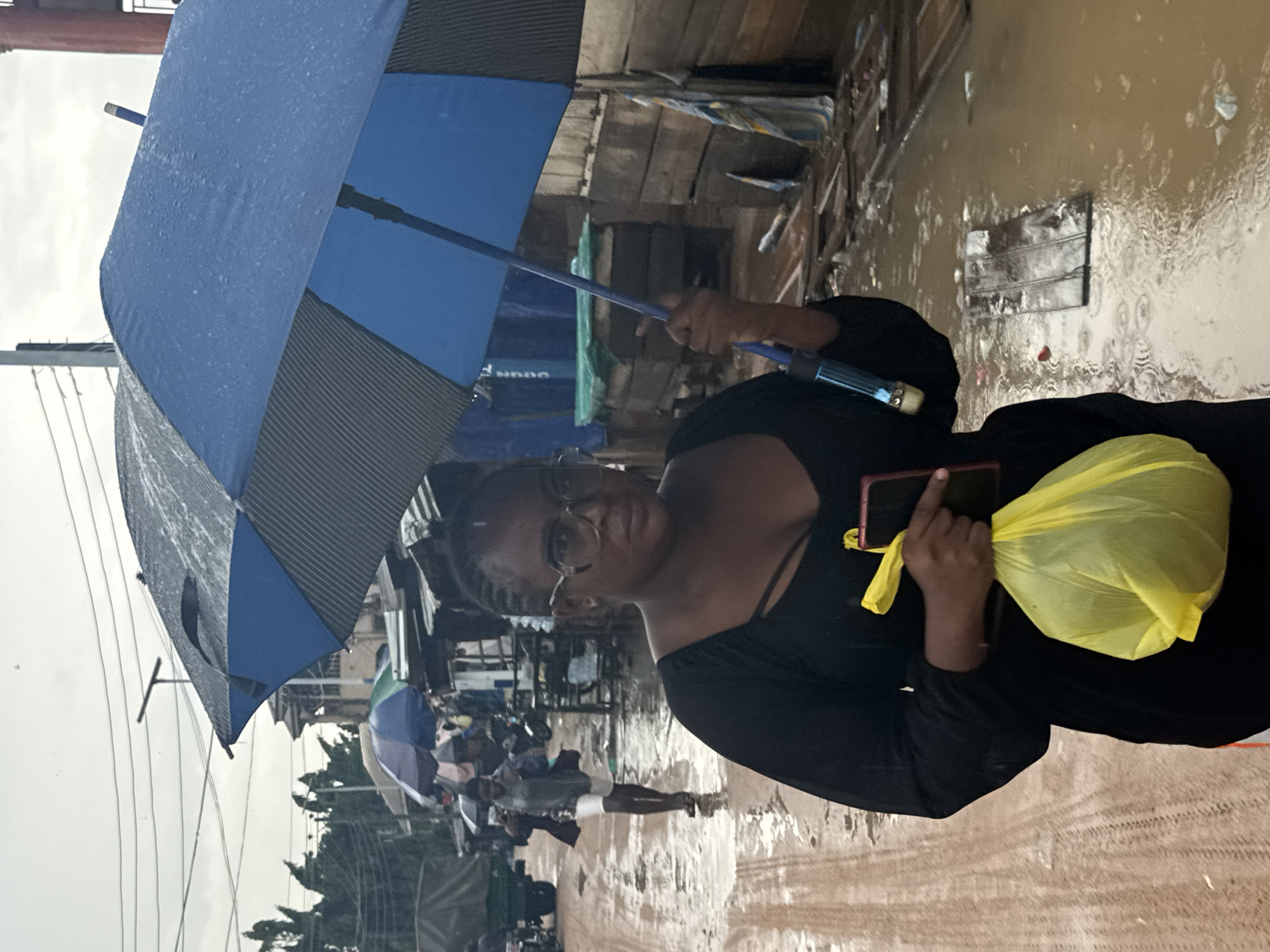 The image captures a person navigating a flooded street in Ihiagwa, Nigeria, during what appears to be heavy rain. The main subject, positioned prominently in the foreground, is a person with dark skin, wearing glasses and a black long-sleeved top. They hold a blue and dark grey umbrella over their head, which is visibly wet with raindrops. In their left hand, they hold a smartphone, and in their right hand, they carry a bright yellow plastic bag. Their expression is neutral as they look towards the camera.
The setting is a busy street or pathway, heavily inundated with muddy, brown floodwater. Ripples and reflections on the water's surface suggest ongoing rain and movement. In the background, other individuals, some also holding umbrellas, can be seen attempting to traverse the waterlogged area. Various structures, possibly informal market stalls or residential buildings, line the street, many appearing to be constructed from wood and corrugated metal. The overall atmosphere indicates challenging weather conditions and daily life continuing despite the flooding. The sky is overcast, contributing to the dim lighting. No clearly discernible text is visible on any objects or signs within the frame.