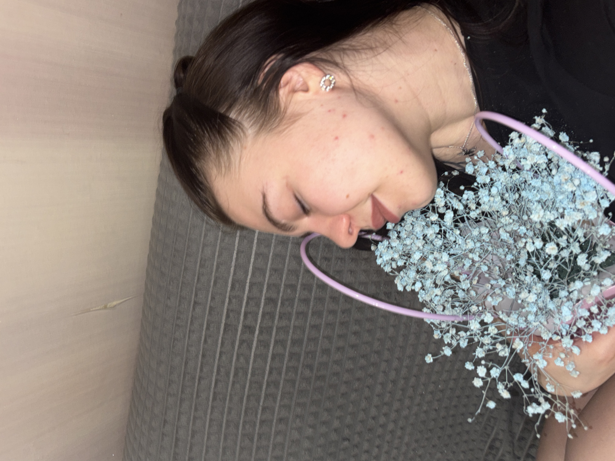 A young woman lies on a gray, horizontally-striped surface, possibly a bed or couch, while holding a bouquet of small, light blue flowers. Her face is visible, showing closed eyes, a silver stud earring in her left ear, and a silver necklace around her neck. She has fair skin with some blemishes. She wears a black shirt. The location in the city of Perm, Russia, cannot be determined from the image.