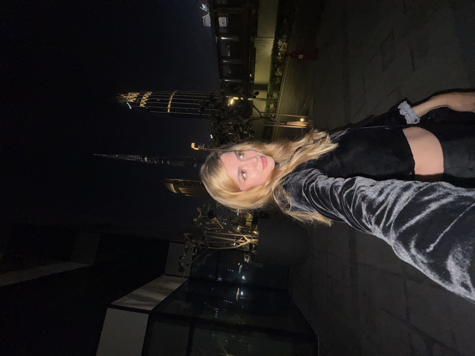 A woman with blonde hair takes a selfie at night, with Dubai's skyline in the background. She wears a dark, velvety long-sleeved top and matching bottoms, with a visible midriff. The perspective is slightly tilted, emphasizing the city's illuminated buildings, including the Burj Khalifa, against the dark night sky. A planter is located on her right.