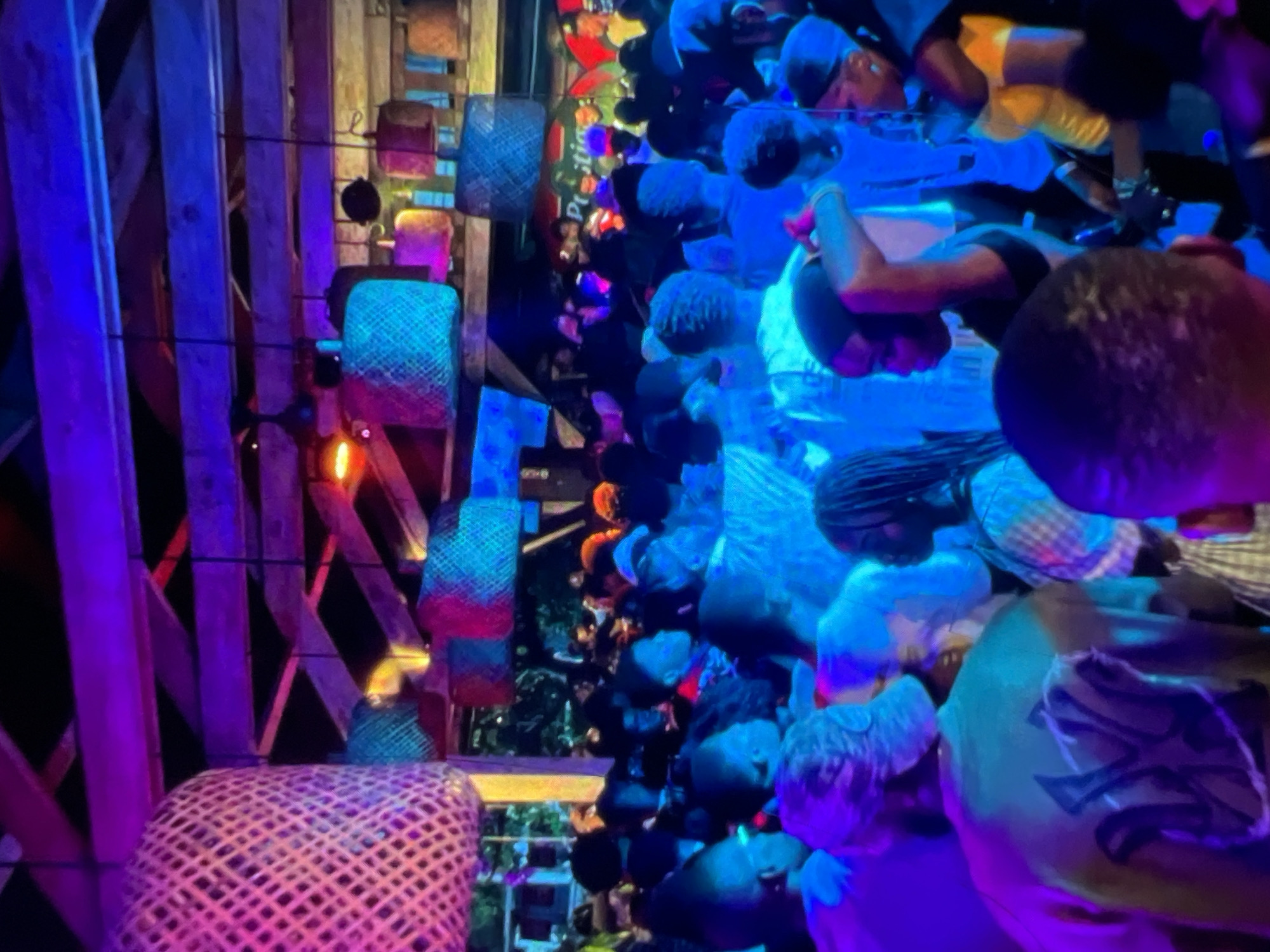 The image depicts a crowded indoor venue, likely a club or bar, illuminated by vibrant purple and blue lights. The ceiling is adorned with woven lampshades and exposed wooden beams, suggesting a rustic or tropical aesthetic. A diverse group of people, predominantly young adults, are visible, some dancing, others conversing, and a few observing. The atmosphere appears lively and festive, with the presence of music and social interaction. The text "Prestige" is partially visible on a sign in the background, possibly indicating the name of the establishment or an event. While the specific location in Lagos, Nigeria cannot be definitively determined from the visual information alone, the scene is consistent with nightlife venues found in urban settings.