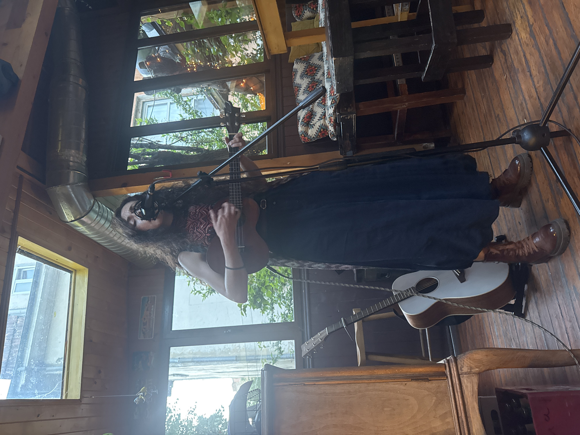 The image captures a female musician performing indoors during what appears to be daytime, likely in a cafe or small venue in Sofia, Bulgaria. The central figure is a woman with long, dark, curly hair and glasses, positioned in front of a microphone on a stand. She is wearing a dark top with a red, black, and white geometric pattern around the neckline, a long dark blue skirt, and sturdy brown lace-up boots. Her hands are engaged in playing a small stringed instrument, possibly a ukulele or mandolin.
The setting features warm, wooden interiors, including paneled walls, floorboards, and ceiling beams. Large windows line the background, letting in abundant natural light and showcasing green foliage from trees or bushes outside, along with glimpses of other buildings. A prominent corrugated metal air duct runs along an upper wall, and a patterned cushion with a design similar to the performer's top is visible on a surface in the background. An acoustic guitar with a white top and natural wood body rests on a stand behind the performer, and a wooden cabinet or counter is also part of the furniture. The overall atmosphere suggests a live, intimate musical performance. No discernible text is visible in the image.