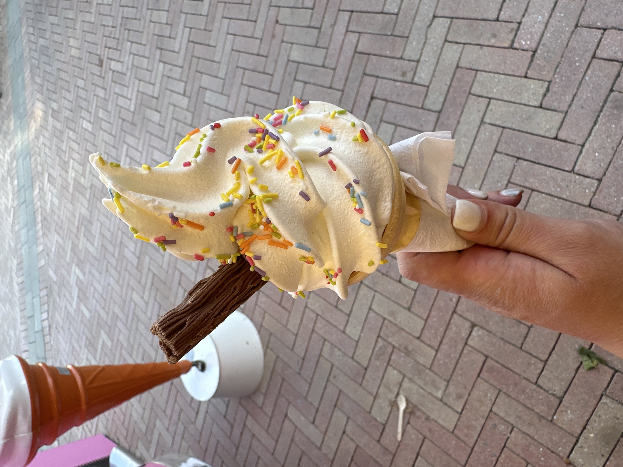 A high-angle shot features a hand holding an ice cream cone adorned with colorful sprinkles. The ice cream is swirled and attached to a chocolate stick. A white paper napkin partially covers the hand holding the cone. In the background, an orange-colored cone sits in a white holder. The setting appears to be outdoors, with a brick-patterned pavement serving as the backdrop. The location details "Unknown, Unknown" suggest the specific city is unknown.