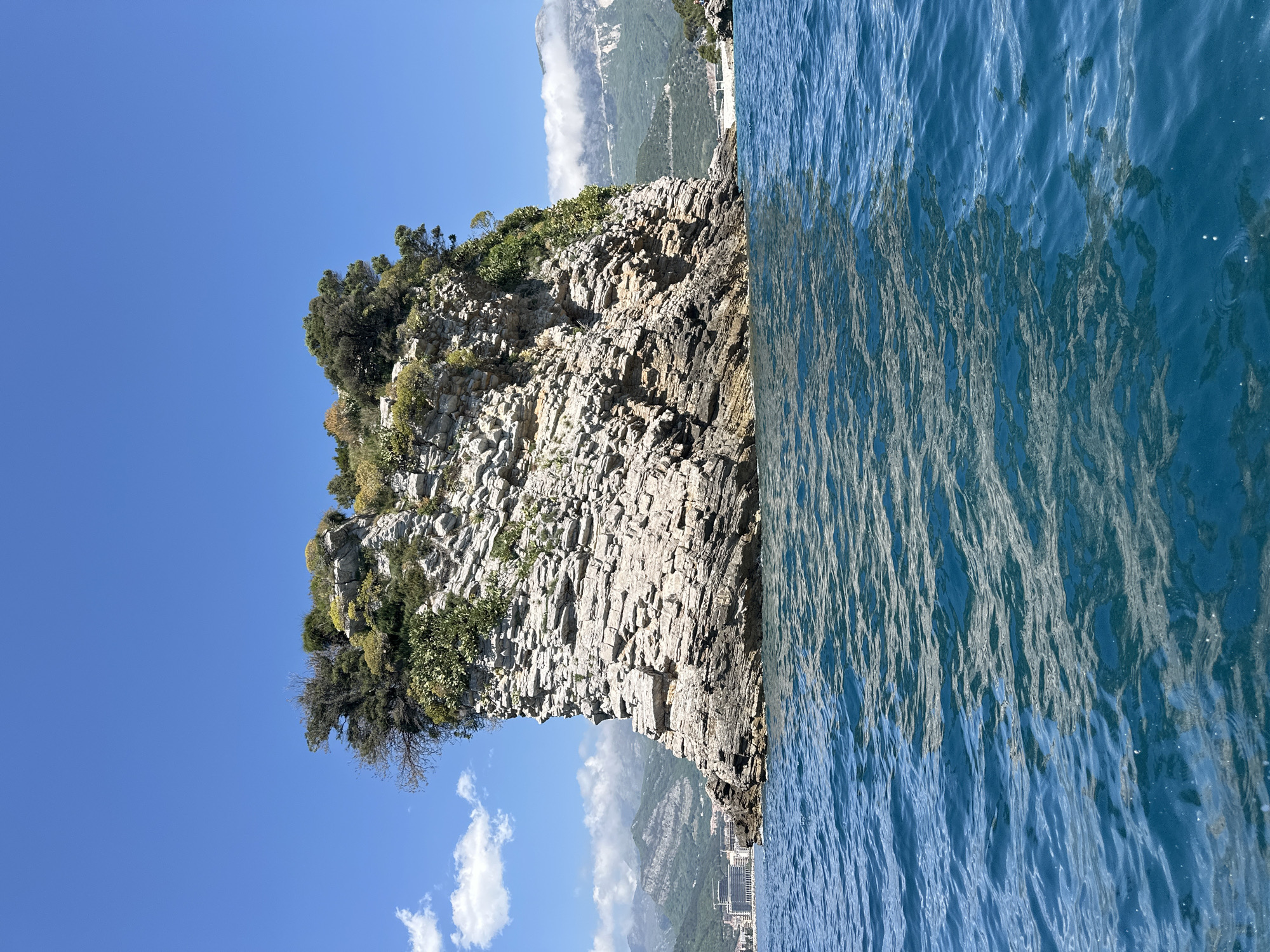 The image depicts a rocky cliff extending into the sea, with a blue sky and white clouds above, a mountain in the background, and the city of Budva, Montenegro, visible on the coastline. The sea appears blue and rippled. The cliff face is layered with vegetation on top.