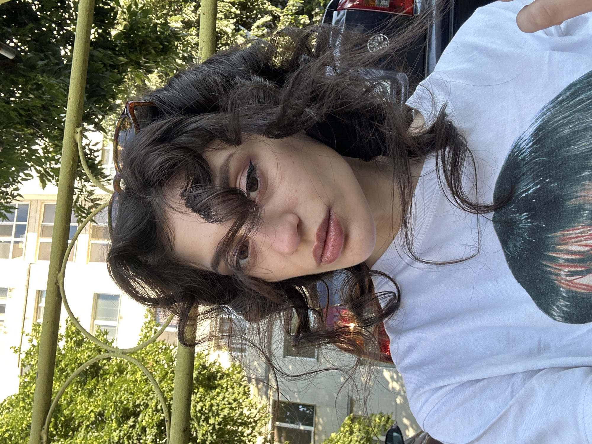 A young woman with dark, curly hair, wearing a white t-shirt, poses for a selfie outdoors. Her hair is styled in large curls and is partially obscuring her face. She is wearing light makeup, including eyeliner and lip gloss. In the background, a green metal fence is visible, with trees and a building behind it. A dark-colored car is parked in the background. The lighting suggests it is daytime. The image is a close-up of the woman's upper body and face. It is not possible to determine the specific location in the city of Sofia, Bulgaria from the visual information provided.