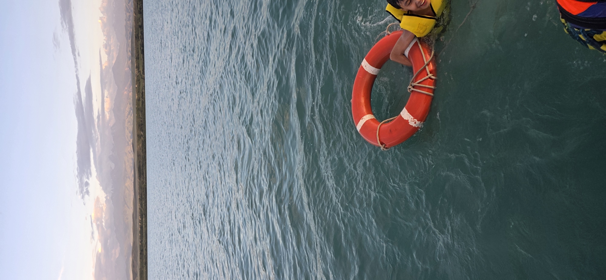 The image depicts a young boy wearing a yellow life jacket, smiling as he holds onto a bright red life preserver in the water. The boy's legs are visible as he floats. The water is a deep teal color with visible ripples and waves, suggesting it is a natural body of water. In the background, a coastline with mountains and a soft, cloudy sky can be seen. The sky suggests it might be sunset or sunrise due to the muted colors and soft light. The overall scene conveys a sense of joy and perhaps a moment of recreation or a lesson in water safety. There is no visible text in the image.