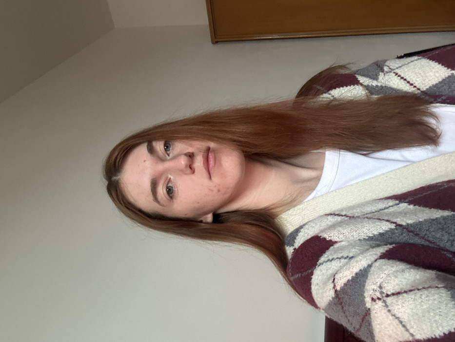 A fair-skinned person with long, straight, reddish-brown hair is facing the camera. They are wearing a white t-shirt and a burgundy, grey, and white argyle cardigan. The background consists of a light grey wall and a dark wooden cabinet in the top right corner. The image has a slightly tilted perspective. The person's expression is neutral.