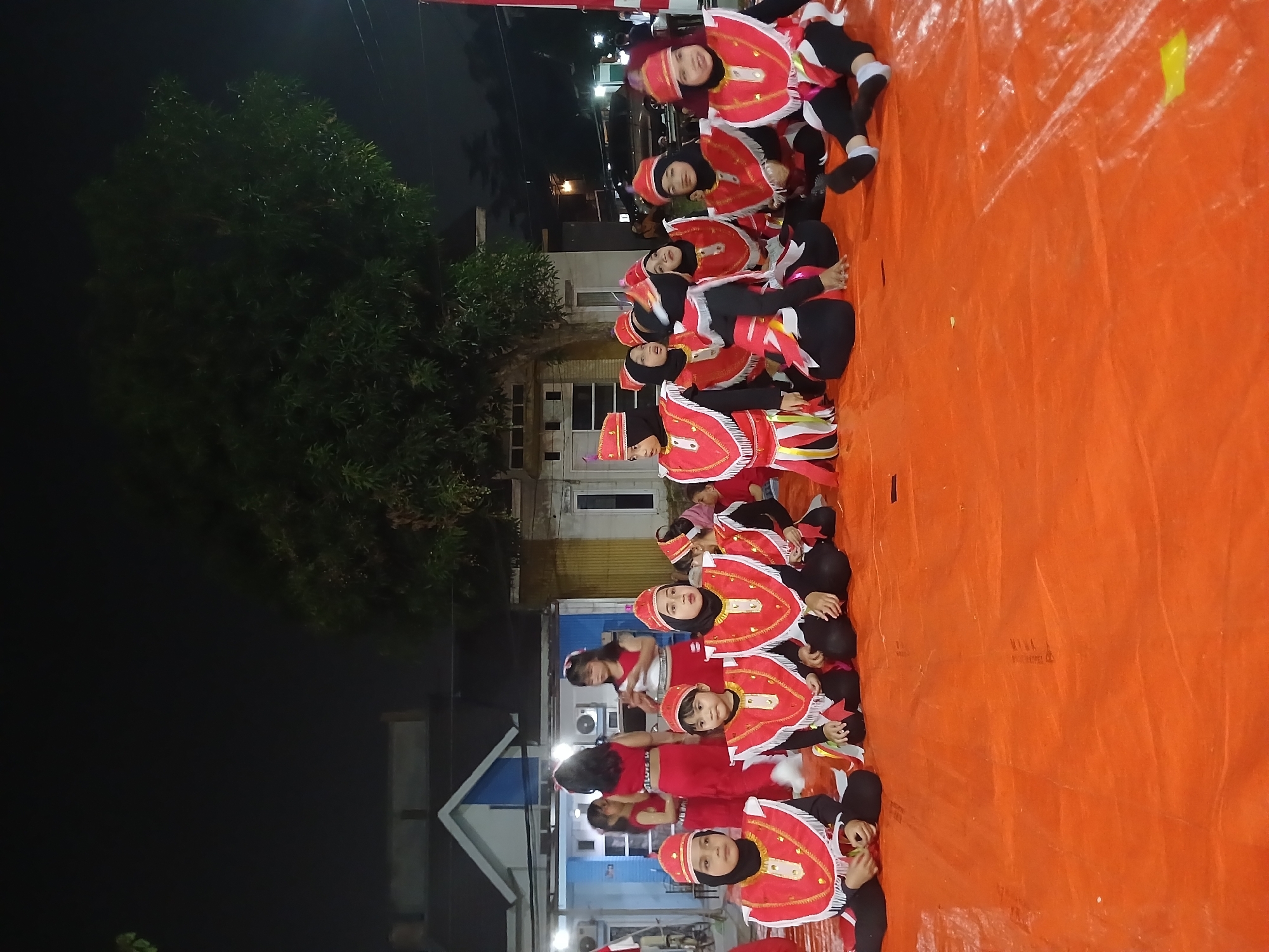 In Kebalen, Indonesia, a group of young girls dressed in traditional red and white costumes, complete with headwear and white trim on their vests, are seated on an orange tarp. They appear to be participating in a performance or cultural event outdoors at night, indicated by the dark sky and visible tree branches. Behind them, a building with a blue and white facade and air conditioning units is visible. The scene is illuminated by artificial lights, suggesting a stage or designated performance area. The girls are positioned in rows, some looking towards the viewer, others engaged with their surroundings. The overall atmosphere is one of a community gathering or celebration.