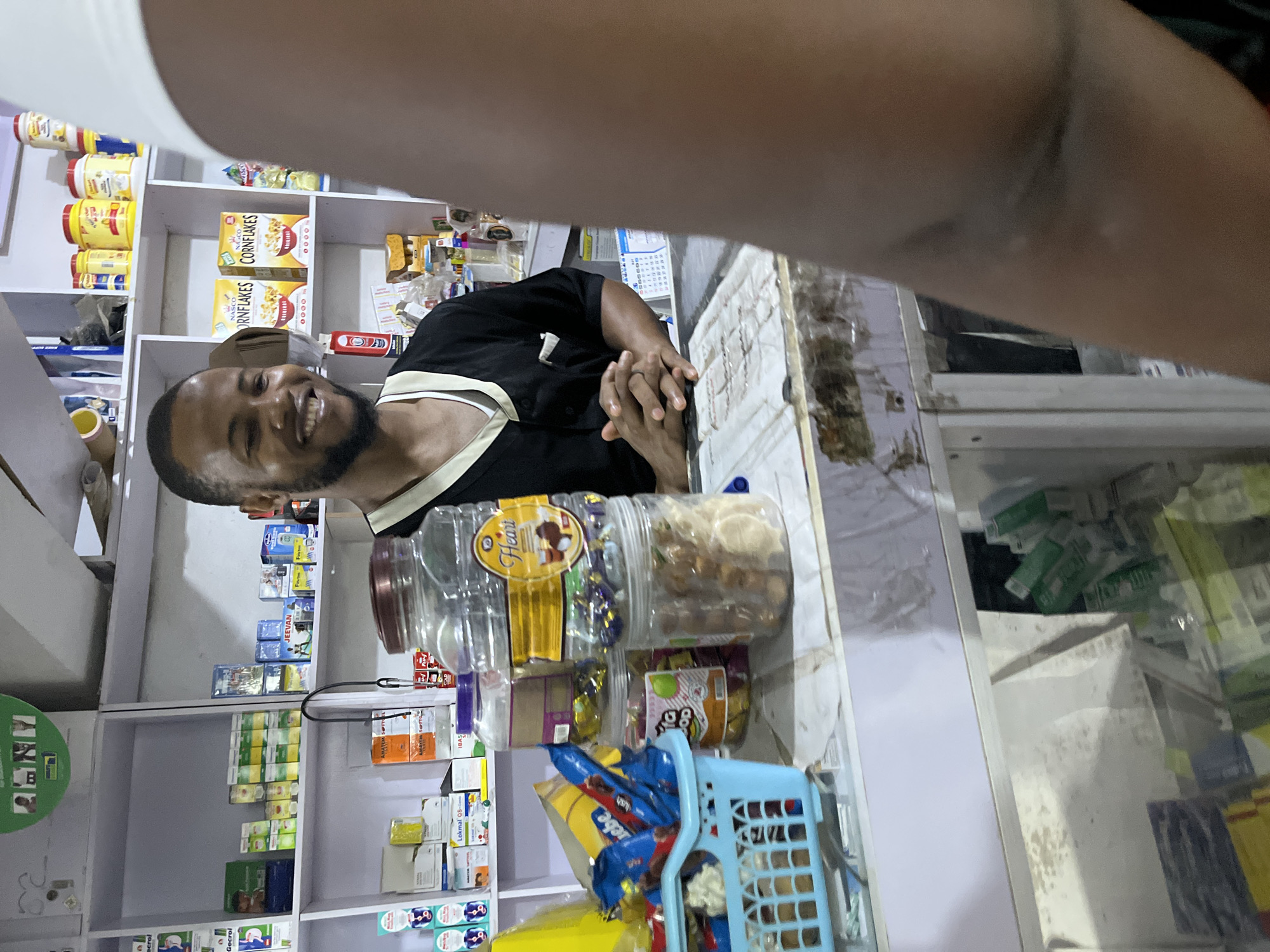 A man stands behind a counter filled with goods in a store, likely in Abuja, Nigeria. The store shelves are stocked with various products, including cereals, candies in plastic jars, and medication boxes. The man, who has a beard, is smiling and looking toward the viewer, with his hands clasped together on the counter. The store appears to be a small, local business.