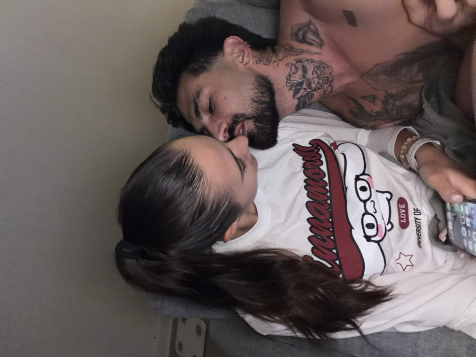 The image displays two individuals, a female and a male, engaged in close interaction within an indoor environment. The female has long, dark hair secured in a high ponytail and wears a white long-sleeved shirt with a "Cinnamoroll" graphic and "University of" text. A light-colored beaded bracelet is visible on her right wrist. She holds a smartphone displaying an app grid in her left hand. The male, shirtless, exhibits a trimmed dark beard and mustache, along with multiple tattoos on his neck and upper chest, including a prominent dual-face mask design and a stylized mouth. He has an earring in his left ear. Their faces are positioned very close, nearly touching at the lips and nose, indicating an intimate moment. They are situated on what appears to be a soft, light grey seating surface. A white rectangular object is partially visible on a surface in the upper left background. The lighting is subdued. The scene is set in Bangkok, Thailand.