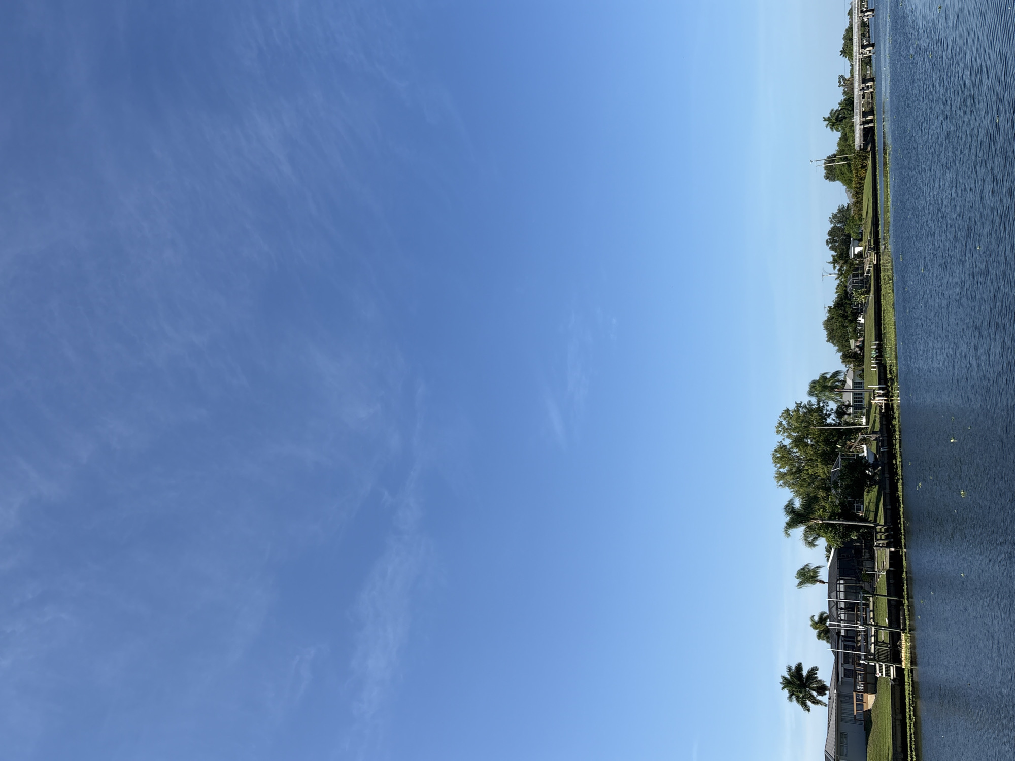 This media file captures a scenic view of a canal in North Port, USA, under a clear blue sky. The image is oriented vertically, with the sky dominating the left side and the water and residential area on the right. Wispy clouds are visible in the upper portion of the sky, suggesting a pleasant, possibly late morning or early afternoon.
The right side of the image showcases a well-maintained waterfront with several houses and their docks. Lush green trees and palm trees line the shore, interspersed with manicured lawns. Some of the houses appear to have screened-in porches or pools, typical of a warm climate. Boats are docked along the piers, indicating recreational use of the canal. A bridge is visible in the distance, spanning the canal. The water itself is dark blue and appears calm, with a narrow band of green aquatic plants along the edge of the canal.
The overall impression is one of tranquility and a peaceful residential setting, likely in a community that values waterfront living and outdoor activities. There is no text visible in the media file.