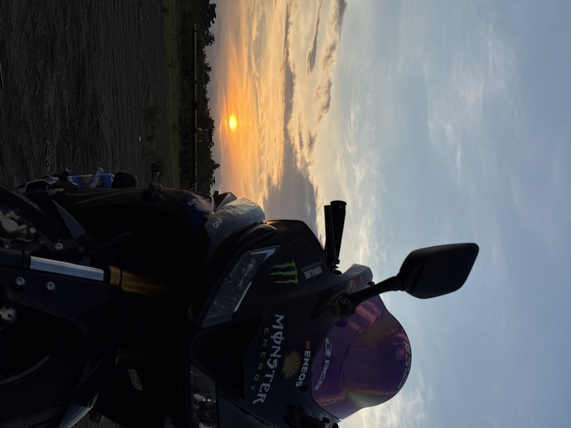 A motorcycle is parked in an open, unpaved area with a sunset in the background. The sky is filled with clouds in shades of orange, yellow, and blue. The sun is visible as a bright orange orb near the horizon. In the middle ground, there is a flatbed trailer and vegetation, including trees. The foreground is dominated by the motorcycle, which is dark blue and black with gold-colored suspension forks. It features logos such as "Monster Energy" and "Eneos". The motorcycle is positioned in a way that its front is closest to the camera, and the background is slightly blurred. There are no people visible in the image. The scene appears to be a rural or undeveloped area within or near Davao City, Philippines, possibly an industrial or storage yard, given the presence of the trailer and the unpaved ground.