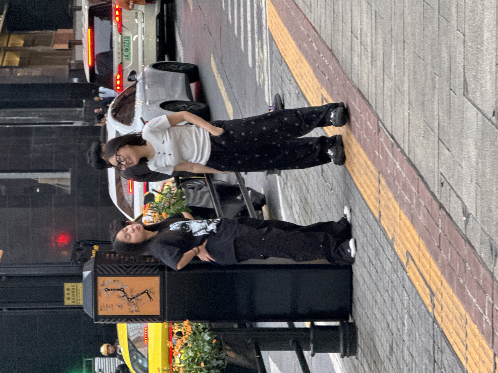 In Yuzhong District, China, two young women stand on a sidewalk next to a street. The woman on the left, wearing a black t-shirt and black baggy pants, has her hands on her hips. The woman on the right, in a white t-shirt and black patterned pants, has one hand in her pocket and is looking to her left. Both are wearing dark shoes. In the background, several cars are visible, including a white sedan and a white SUV with a license plate that reads "AAK2210". A yellow taxi is also present. A black utility pole with a map and a yellow sign is located to the left of the women. The sidewalk is paved with gray bricks and features a raised tactile paving system with yellow and red stripes. The scene appears to be in an urban environment.