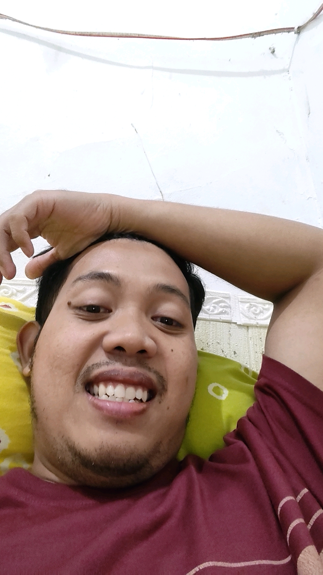 A man is lying on his back, resting his head on a pillow. He is looking at the camera and smiling. He is wearing a maroon t-shirt. The background is a white wall. The image appears to be taken indoors, possibly in a bedroom. There are no visible signs of the location being in Mekarmukti, Indonesia.
