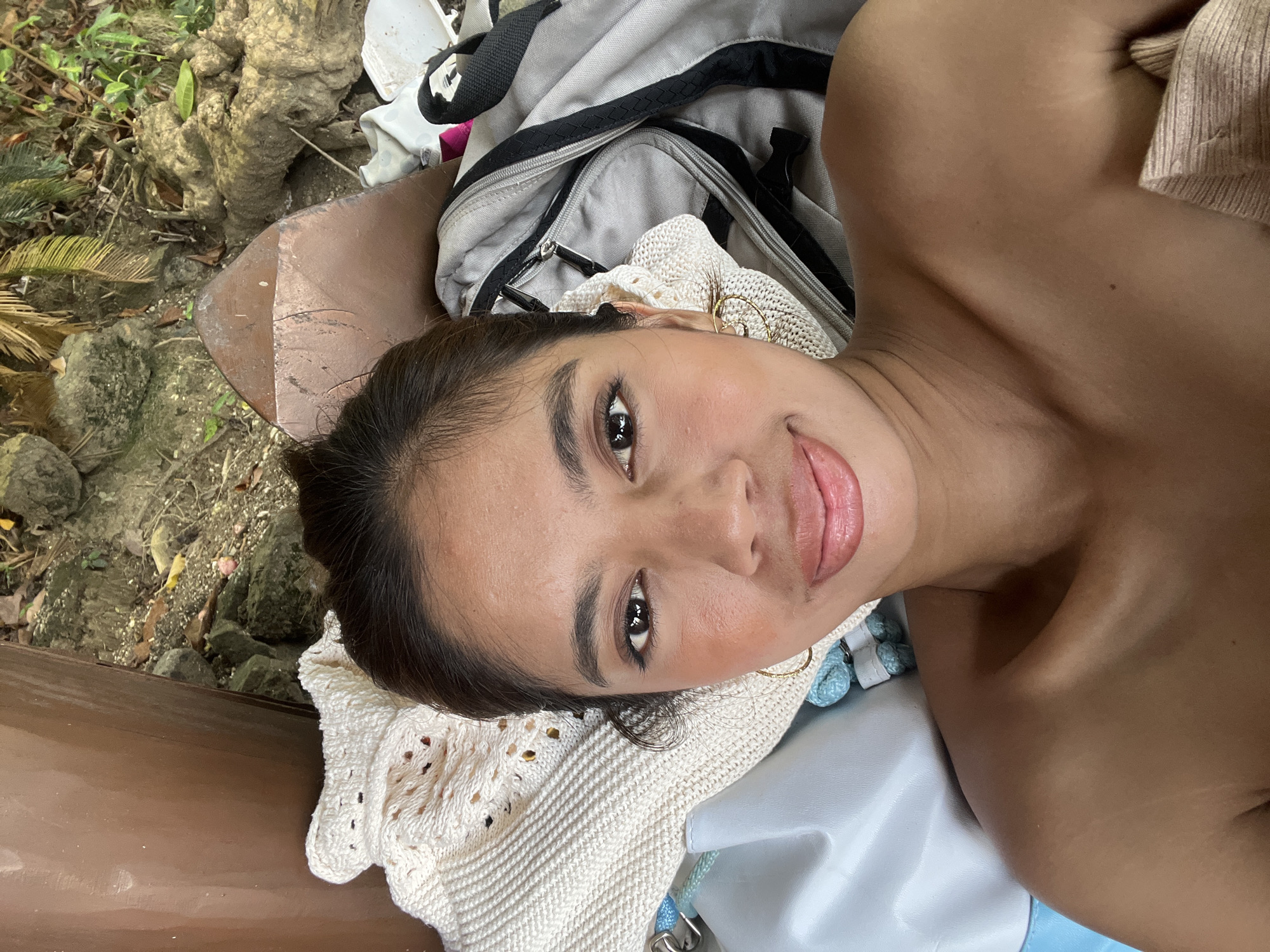 A medium, eye-level, selfie-style shot shows a woman resting in what appears to be a natural environment. She is lying on a white crocheted blanket atop a pale blue bag with light blue rope-style handles, which rests on a light gray backpack. She is fair-skinned with dark hair pulled back and gold hoop earrings. The woman's left side is resting on what appears to be a light brown, wooden beam. Behind the wooden beam is greenery and a tree trunk. The location is identified as the city of Banaybanay, Philippines.