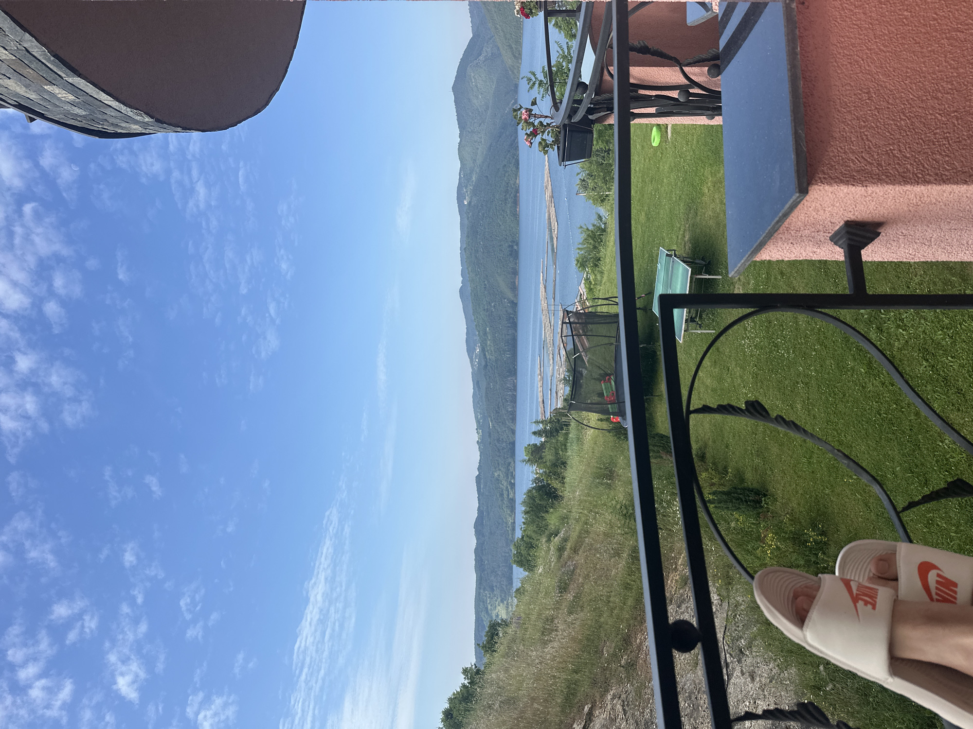 The image presents a first-person perspective from a balcony in Glavatartsi, Bulgaria, overlooking a vast reservoir surrounded by verdant hills under a clear blue sky with scattered white clouds.
In the immediate foreground, a person's lower legs and feet, adorned with white Nike slide sandals featuring an orange logo, are visible, resting casually near a black wrought-iron balcony railing. The railing, characterized by its sturdy bars and decorative scrollwork, extends across the lower portion of the frame. To the right, a segment of the building's exterior, specifically a reddish-pink, textured concrete pillar topped with a dark, possibly granite or slate, slab, is visible. Black planters with blooming pink and red flowers are attached to the balcony railing on the right. A faint crescent moon is subtly visible in the upper right quadrant of the sky.
The midground features a well-maintained green lawn directly below the balcony. On the grass, a large trampoline with a protective netting enclosure is situated on the left, and a green outdoor ping-pong table with a white net is centrally placed. A bright green plastic ring lies on the grass near the right edge of the lawn. Beyond the lawn, a dry, sloping hillside with sparse vegetation and some exposed earth descends towards the water on the left.
The background is dominated by a large, calm blue body of water, likely the Kardzhali Reservoir, extending across the entire width of the scene. Numerous light-colored, elongated rafts or log formations are clustered on the water, suggesting activities like fish farming or timber storage. Distant, forested hills and mountains, characterized by varying shades of green, rise from the far bank of the reservoir, receding into a lighter atmospheric haze. The sky above is a bright, clear blue, dotted with small, wispy white cumulus clouds.