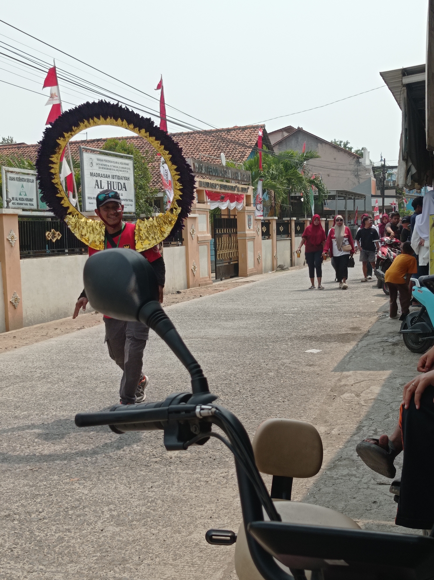 A man in a red shirt and gray pants with a decorative headpiece walks down a street in Kramat, Indonesia. He is wearing a black baseball cap and sunglasses. The headpiece is circular with black and gold decorations and resembles large wings. He is smiling and appears to be participating in a parade or celebration. In the background, there are buildings decorated with red and white flags, indicating a patriotic event. Several people are walking in the street and on the sidewalk, some of whom are also holding drinks. There are also several motorcycles parked on the side of the road. The scene suggests a festive community gathering.