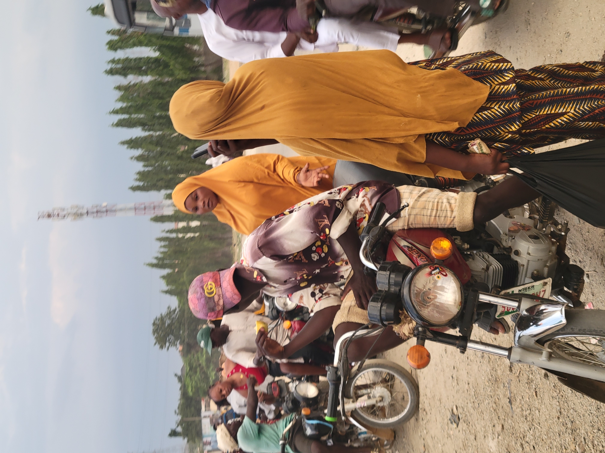 The scene shows a group of people on motorcycles in what appears to be a crowded street in Madalla, Nigeria. Three people are visible on one motorcycle: a driver wearing a cap and a patterned shirt, a woman in a yellow hijab, and a third person wearing a yellow hijab and patterned skirt. Other motorcycles and people are visible in the background, suggesting a traffic jam or gathering. Overhead wires are visible in the upper left, indicating an urban environment.