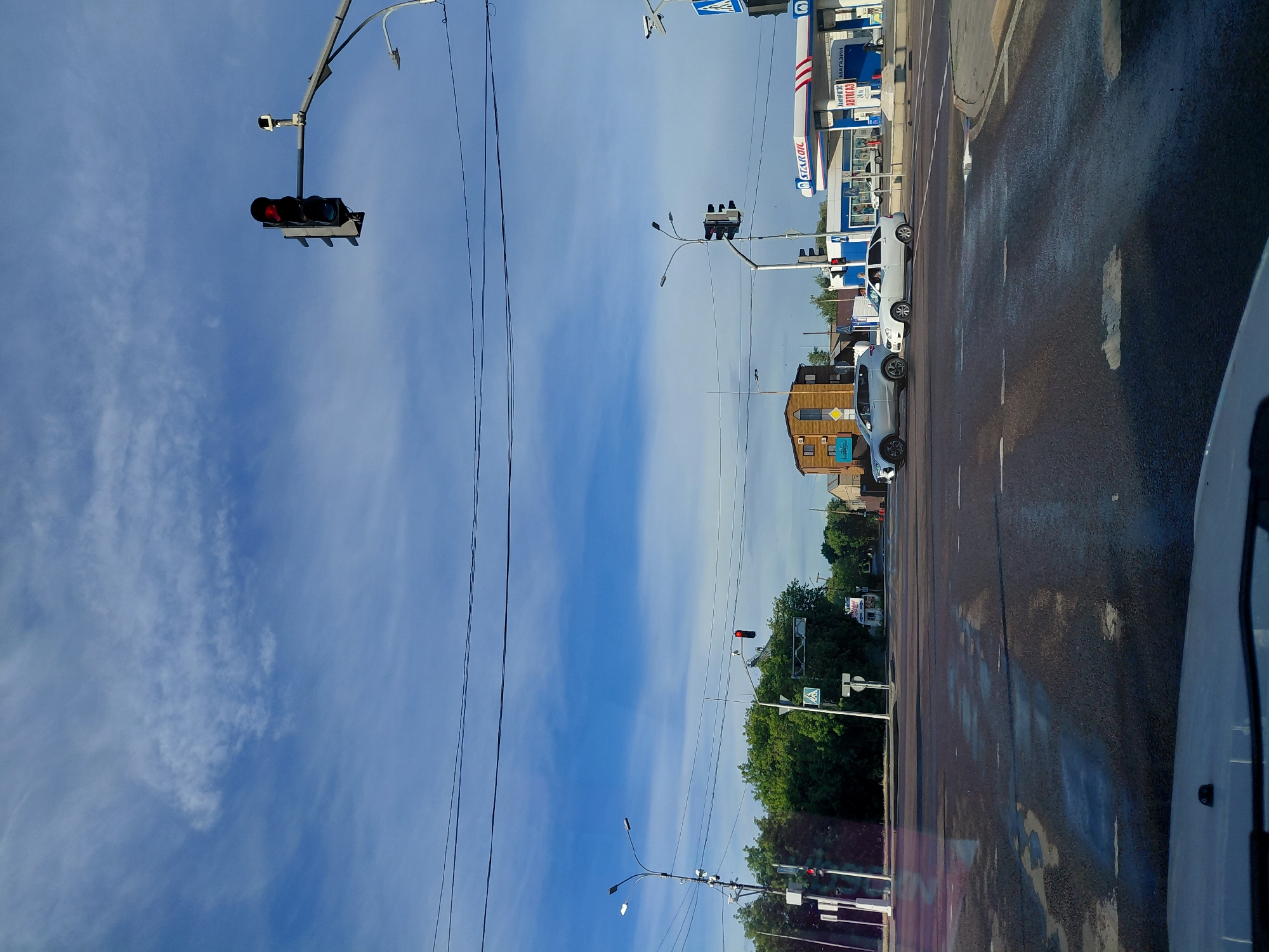 The image depicts a street scene in Qarağandı city administration, Kazakhstan, under a partly cloudy blue sky. A traffic light with a red signal is visible, indicating a stop. Several cars are present at an intersection, with one silver SUV and a white sedan in the foreground. A gas station with the "STAROIL" logo and a blue awning is situated to the right of the intersection. Trees line the background, and various streetlights and power lines are scattered throughout the scene. The road surface appears wet in patches, suggesting recent rain or cleaning. A pedestrian crossing sign is also visible. The perspective suggests the photo was taken from inside a vehicle.