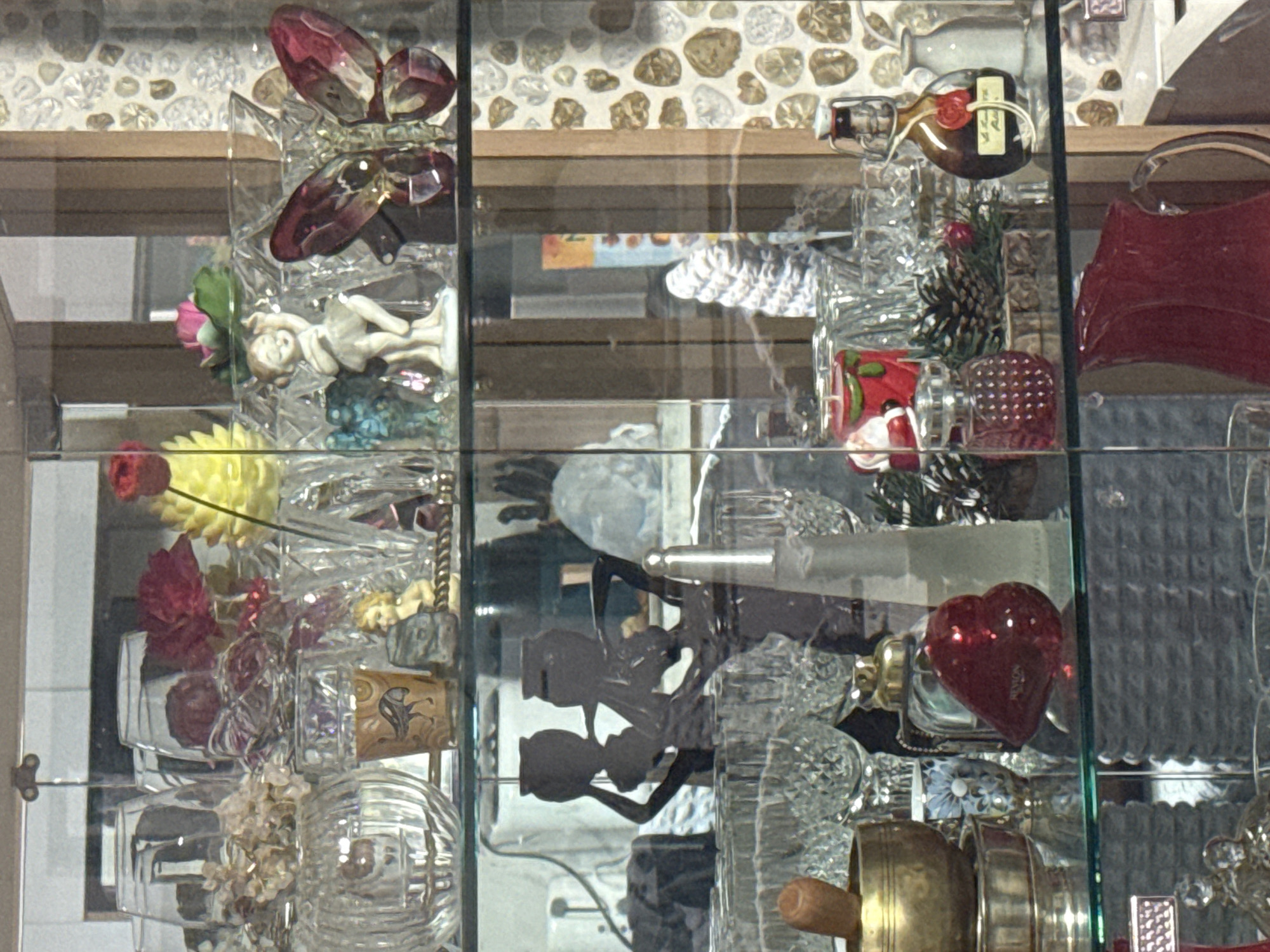 The image shows a glass cabinet filled with various decorative items, predominantly glass and ceramic figurines and ornaments. On the top shelf, there are ornate glass vases, artificial flowers in shades of red, yellow, and pink, and a small figurine of a child. To the right, a large, faceted glass butterfly in deep red and clear glass is displayed.
The middle shelf features a dark silhouette of two figures holding vases above their heads, with a scattering of glassware below. Further to the right, there is a collection of pinecones and what appears to be a red candle in the shape of a Christmas stocking with Santa Claus. A small, dark glass bottle with a cork stopper and a yellow label is also present.
The bottom shelf contains a golden mortar and pestle, a red heart-shaped object with "REVLION" imprinted on it, and a clear glass pitcher with a red liquid, possibly a beverage, situated in the foreground. The background of the cabinet is a light-colored, textured wallpaper.
The image is a close-up shot, and there is no visible indication of its location in Ulaanbaatar, Mongolia. The scene is static, with no discernible actions or interactions occurring.