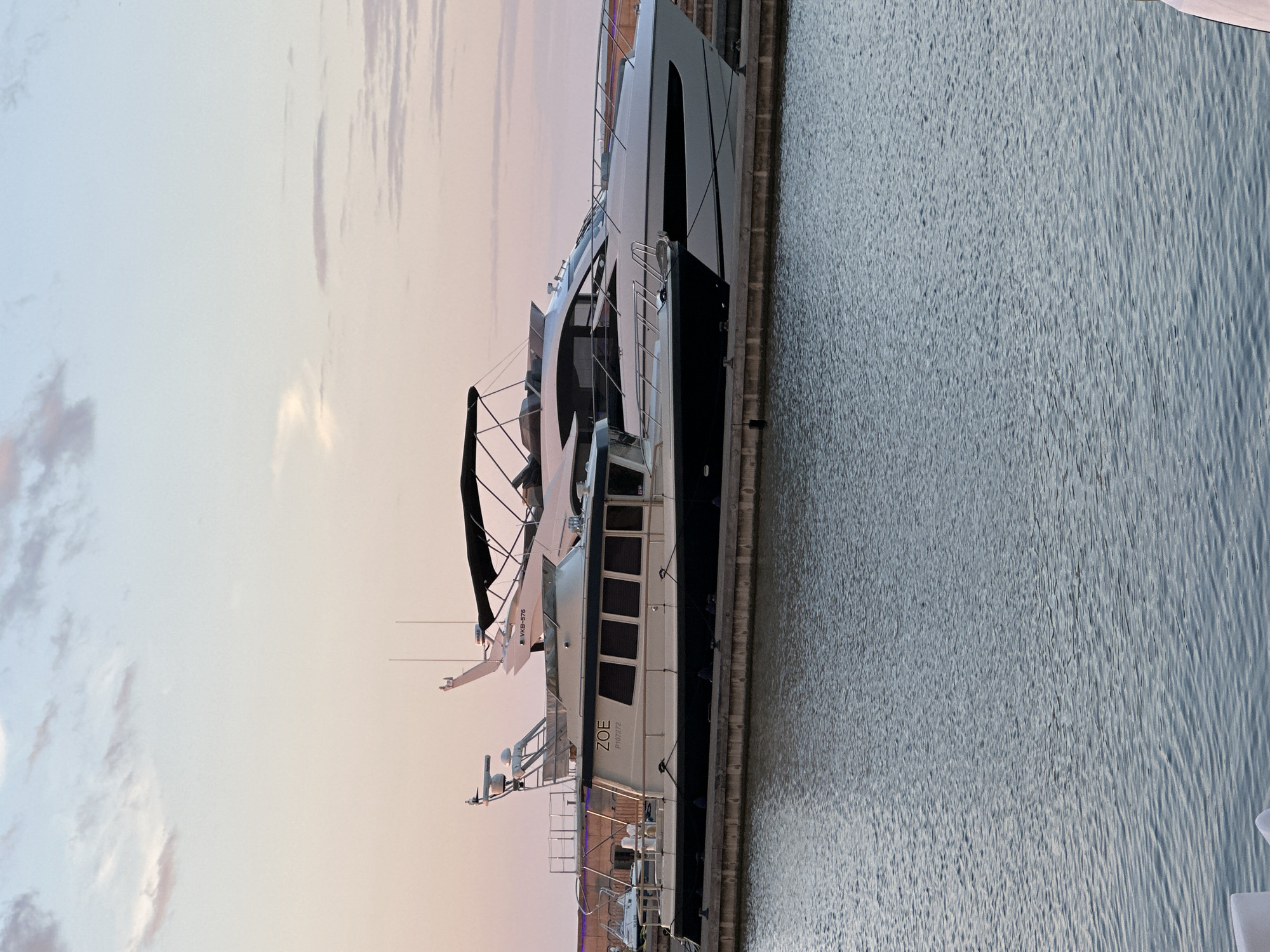 Two yachts are docked side-by-side in a harbor. The yacht in the foreground is a trawler-style boat with a cream-colored hull and a dark blue waterline. It has the name "ZOE" and the registration "P107272" visible on its side. The yacht in the background is a sleek, white motor yacht with a black canopy. The sky is a soft pastel color, indicating either sunrise or sunset. The water in the foreground is calm with gentle ripples. The scene appears to be in a marina within the city of Tallinn, Estonia.