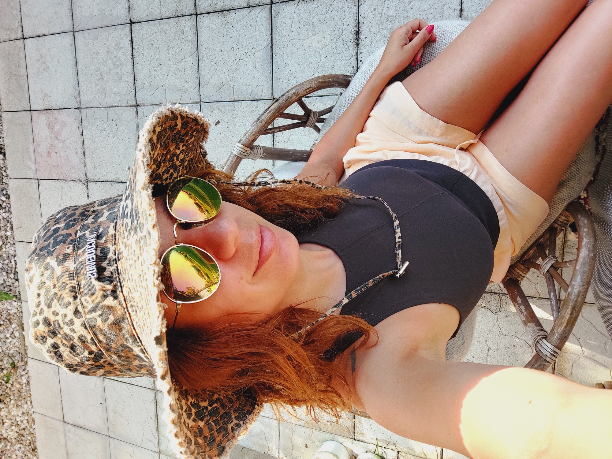 A woman with reddish-brown hair is seated outdoors in a woven chair on a tiled patio. She is wearing a leopard print bucket hat, round sunglasses with reflective lenses, a dark-colored tank top, and light peach-colored shorts. A leopard print cord is draped around her neck and secured to her sunglasses. The setting appears to be a sunny day, with shadows cast by the sunlight. The patio is bordered by gravel and some greenery is visible in the background. The overall scene suggests a relaxed, summery atmosphere. This location is consistent with outdoor areas in Sozopol, Bulgaria, known for its charming patios and outdoor seating.