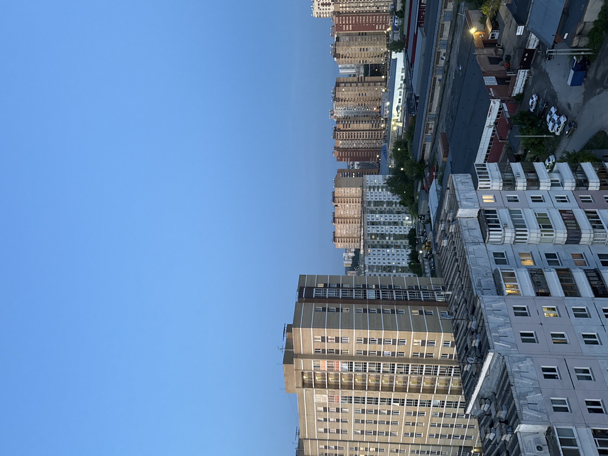 The image displays an urban landscape from a high vantage point, rotated ninety degrees counter-clockwise, with a vast, clear blue sky dominating the left side and city structures filling the right and bottom.
The left portion of the frame is entirely composed of a bright, cloudless blue sky, indicating fair weather. The right and bottom sections are densely populated with buildings. These include numerous multi-story residential apartment blocks, featuring varied architectural styles with repetitive window patterns and visible balconies or loggias. Their facades range in color from light grey and white to beige and brown. Air conditioning units and satellite dishes are visible on many of these buildings.
Below the taller residential structures, particularly in the mid-right area, there are lower-rise industrial or commercial buildings with dark roofs, appearing to be warehouses or workshops. Several light-colored cars are parked in lots or streets within this industrial zone, alongside a large blue dumpster. Scattered green trees are interspersed throughout the urban environment, providing some greenery. Streetlights are visible, with a few already illuminated.
The lighting suggests late afternoon or early evening, characterized by the bright sky combined with lights already on in some building windows and streetlights. The scene is static, with no discernible people or specific activities taking place. No legible text is visible on any of the structures or signs. The architecture is consistent with urban developments found in Russia, aligning with the provided location context of Gamovo.