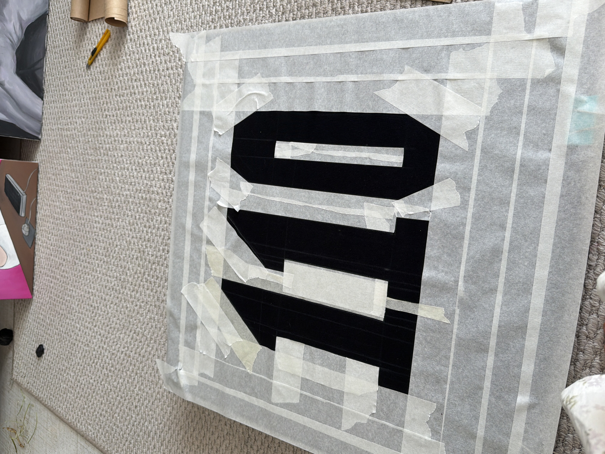 A close-up, top-down view captures a black fabric displaying the number "110" in large, bold font. The fabric is covered by a layer of translucent paper, secured with numerous strips of masking tape. The paper and tape create a border around the number. The scene is set on a light-colored, textured carpet. In the background, a portion of a pink and brown object, possibly a box, is visible on the left, and two rolls of brown paper and a yellow utility knife are on the right. The image does not contain any elements suggesting a specific location in Krasnoyarsk, Russia.