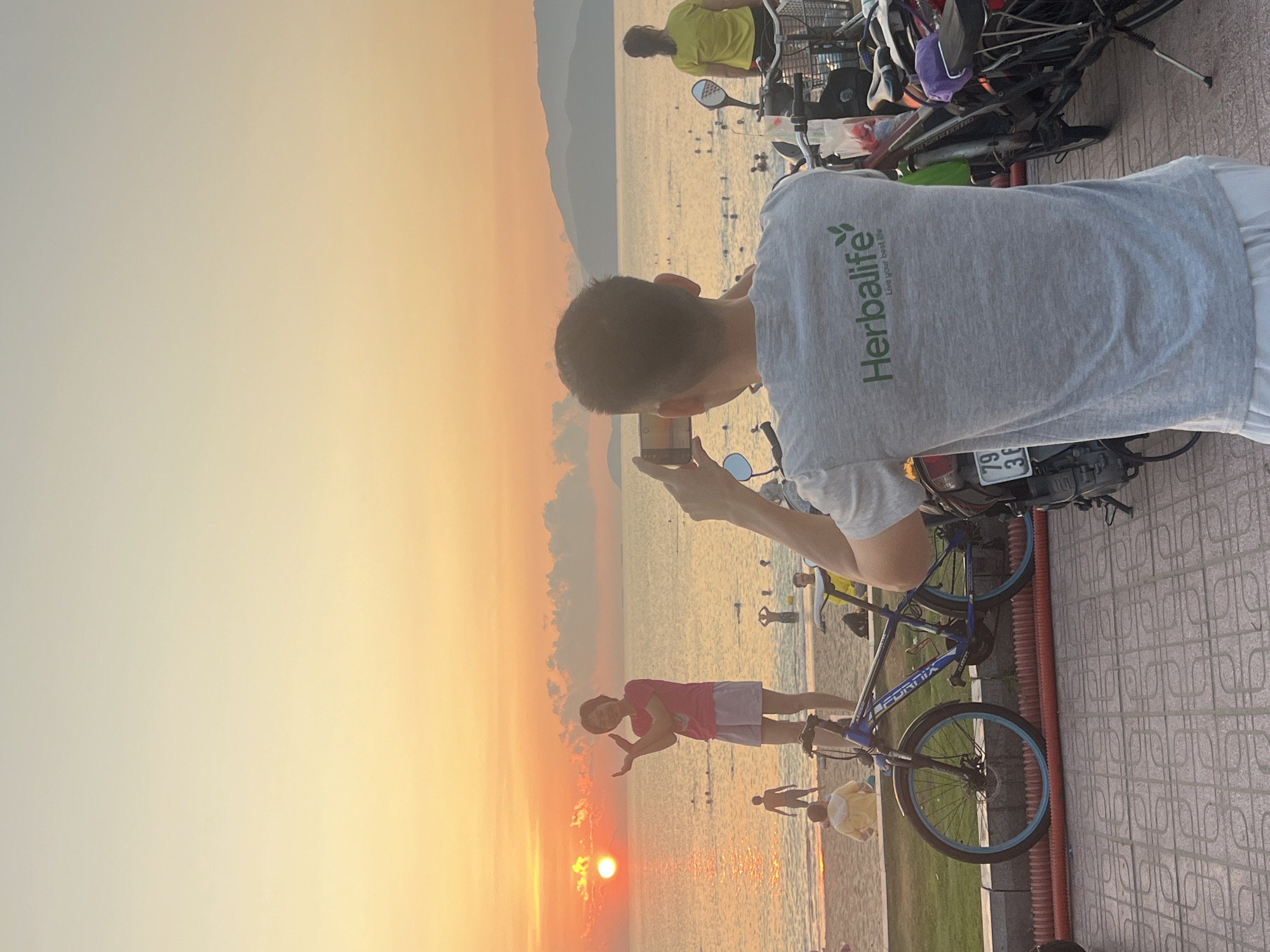 A man in a gray Herbalife shirt is taking a picture with his phone of a sunset at a beach in Nha Trang, Vietnam. A woman in a pink shirt and purple shorts is posing for the camera. There are several people walking on the beach. The man is standing on a tiled pathway with a bicycle and a motorbike parked behind him. The sky is a gradient of orange and yellow.