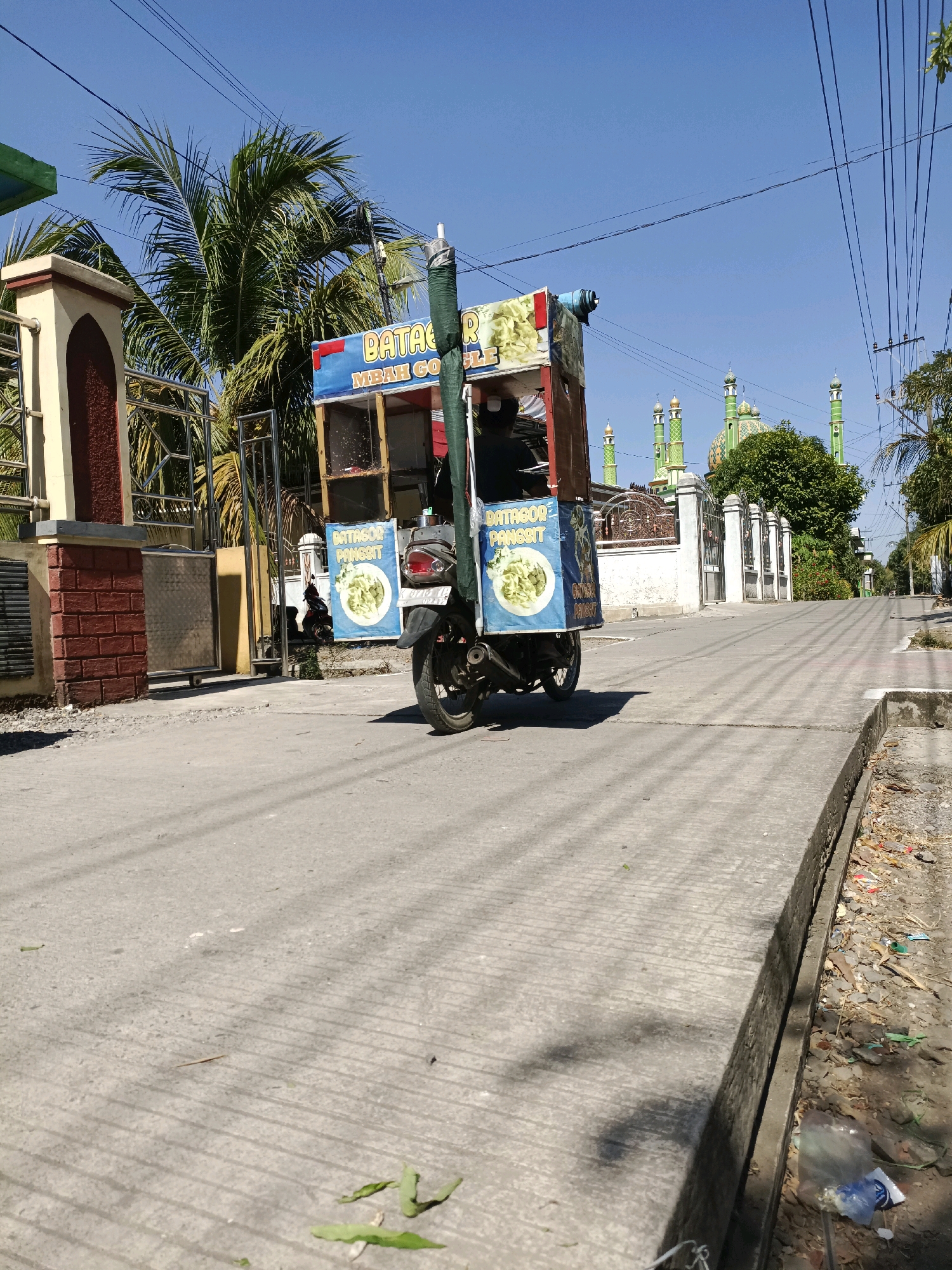This image captures a street scene in Cangkring, Rembang, Indonesia, under a clear blue sky. In the foreground, a paved road is visible, with a raised concrete curb on the right side of the image. The curb area is littered with debris, including plastic bags and dried leaves.
Dominating the mid-ground is a motorcycle that has been modified to serve as a mobile food cart. The cart is brightly colored with blue and red panels, featuring text and images of food. A green tarp hangs from the top of the cart. A portion of a motorcycle's rear tire and exhaust pipe are visible.
In the background, a gate with ornate metalwork leads to a property. A tall palm tree stands behind the gate. Further in the distance, several green-domed mosques with tall minarets punctuate the skyline, suggesting the presence of a significant religious area. The street is lined with power lines stretching across the sky. The overall impression is of a typical, sunny day in a developing urban environment in Indonesia.