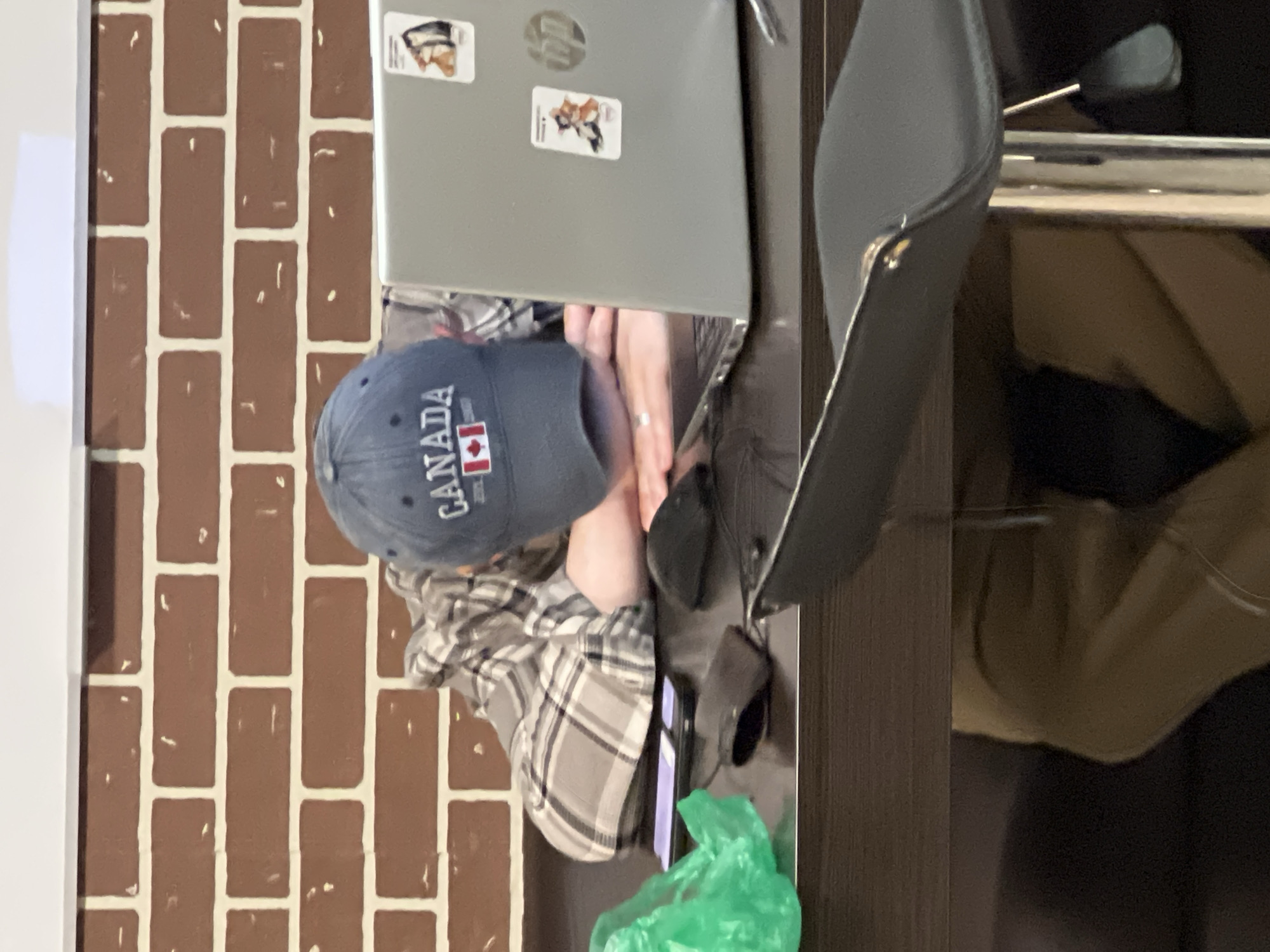 A person wearing a grey and white checkered shirt and a dark blue Canada baseball cap with a red maple leaf is resting their head on a closed grey laptop with stickers. The laptop is on a dark brown wooden desk, next to a black office chair. A black phone and a crumpled green plastic bag are on the desk near the person. The background includes a brick-patterned wall on the left and a blurry dark area on the right. There is no information to confirm or deny if the location is in Khimki, Russia.
