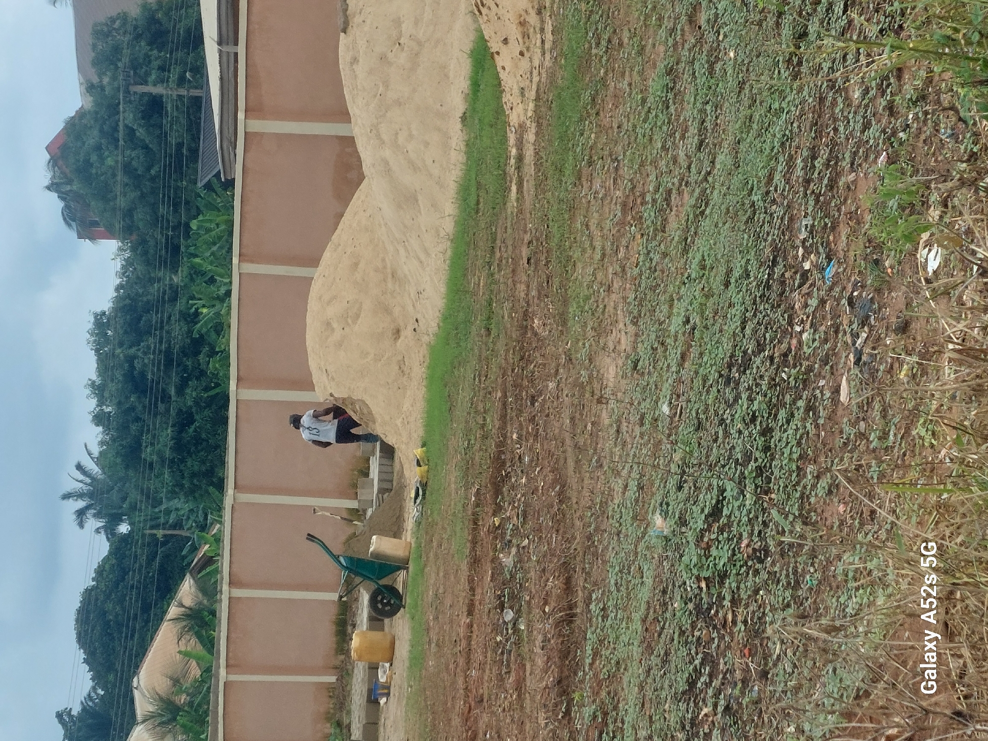 In Umudioka, Nigeria, a person is standing near a large pile of sand next to a tan-colored wall. The person is wearing a white t-shirt with "13" on the back and dark shorts. To the left of the person, a green wheelbarrow is visible, with a yellow container placed next to it. Behind the wall, lush green trees and vegetation are prominent, suggesting a tropical environment. The ground in the foreground is covered with sparse green plants and dry soil, with some scattered debris. The sky appears overcast, indicating a cloudy day. The overall scene depicts a construction or landscaping site. The text "Galaxy A52s 5G" is visible in the bottom right corner of the image, which is a watermark indicating the device used to capture the photo.