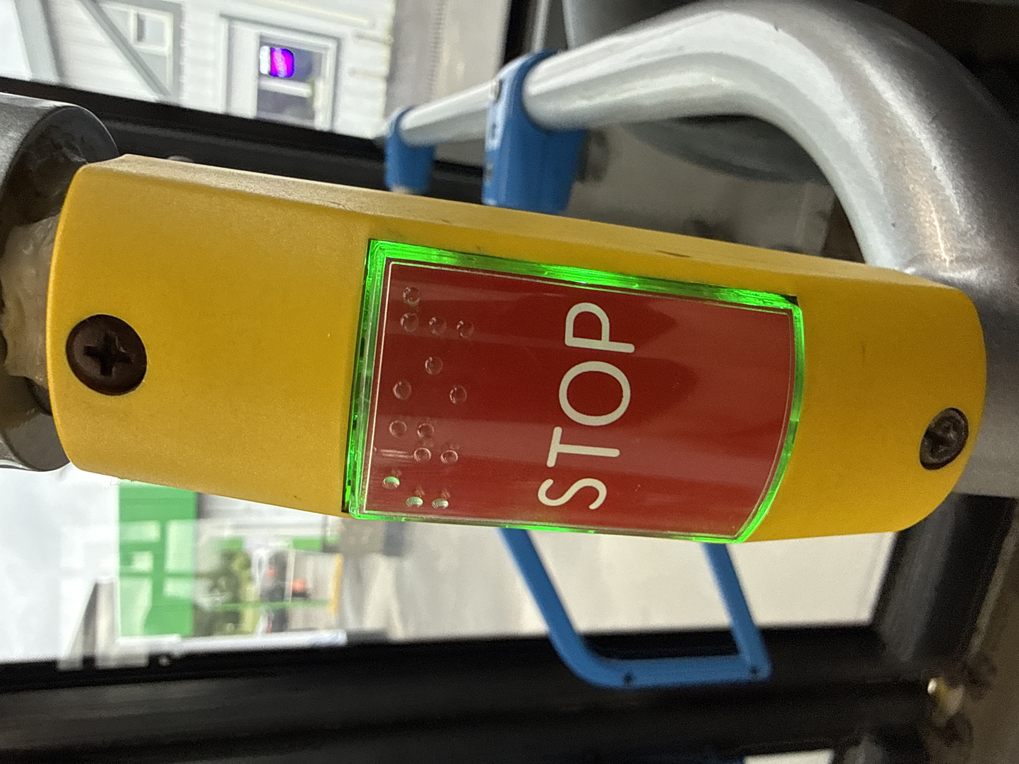 The image shows a close-up of a yellow stop request button on a public transport vehicle, likely a bus, in Yakovlevskoye, Russia. The button is rectangular with a red insert that has the word "STOP" written in white letters. Above the word "STOP" are Braille dots. A bright green LED light outlines the red insert, indicating the button is active. The button is attached to a metallic pole, and a blue grab handle is visible in the background. The environment outside appears to be a street scene with buildings and vehicles visible through the windows. The overall lighting suggests an overcast day. There are no people visible in the image.